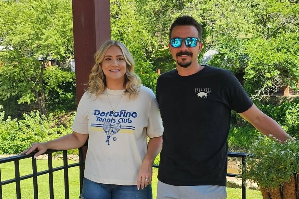 Two people posing on a balcony outdoors; woman in a Portofino Tennis Club shirt, man in a black tee with blue sunglasses, greenery behind.