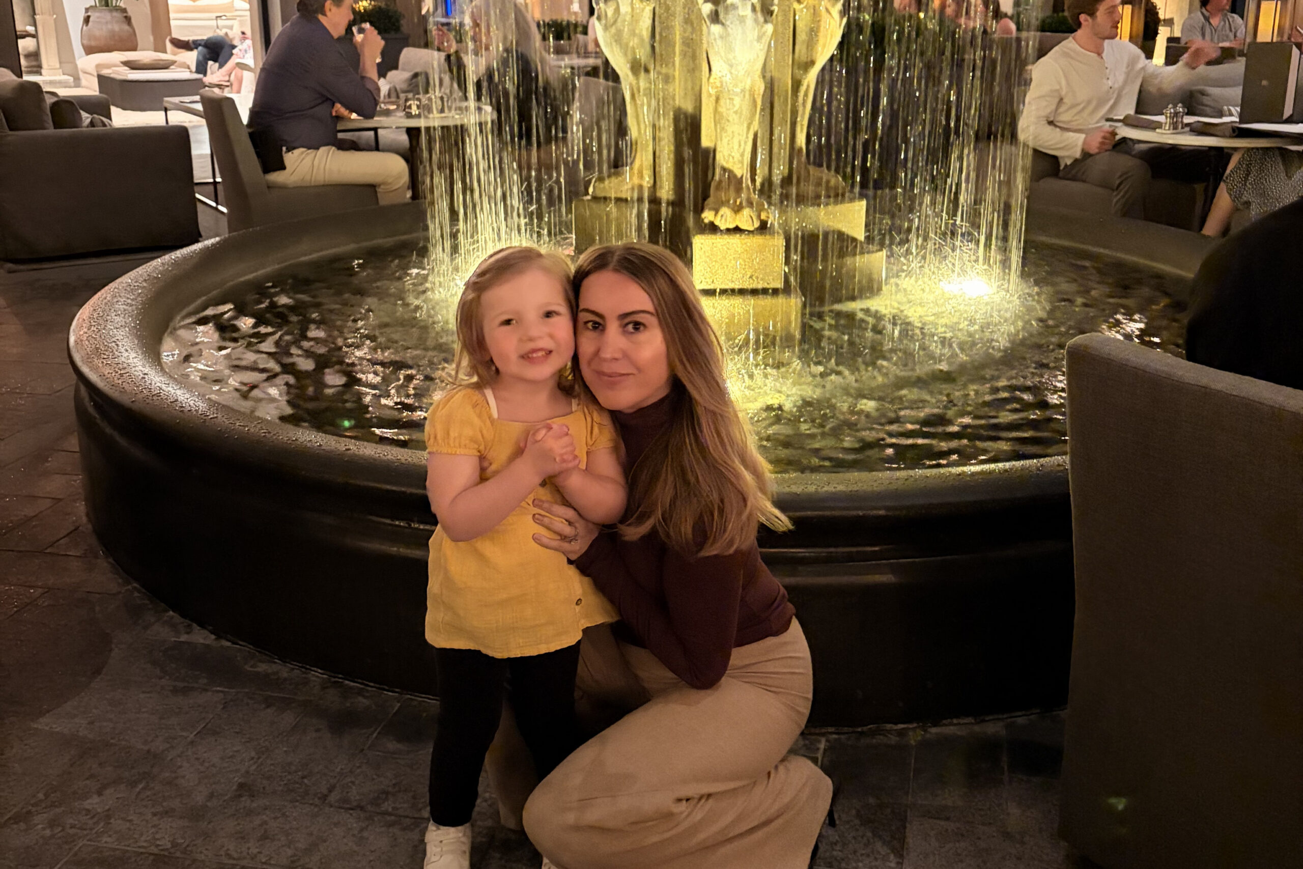 Mother kneels beside her young daughter by a lit fountain in a hotel lobby lounge, posing for a photo.