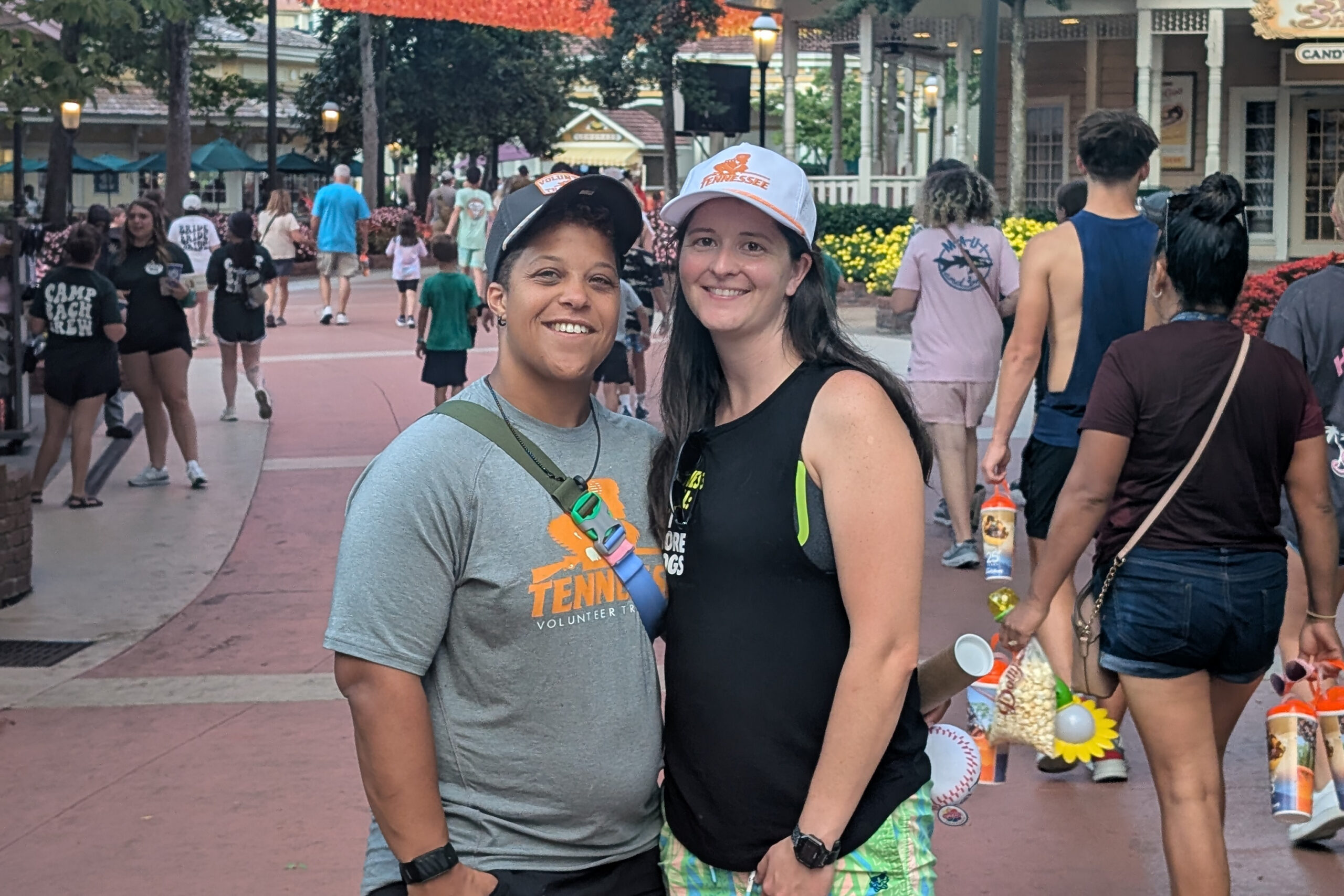 Two women smiling at the camera at an outdoor festival, with a crowd and storefronts in the background.