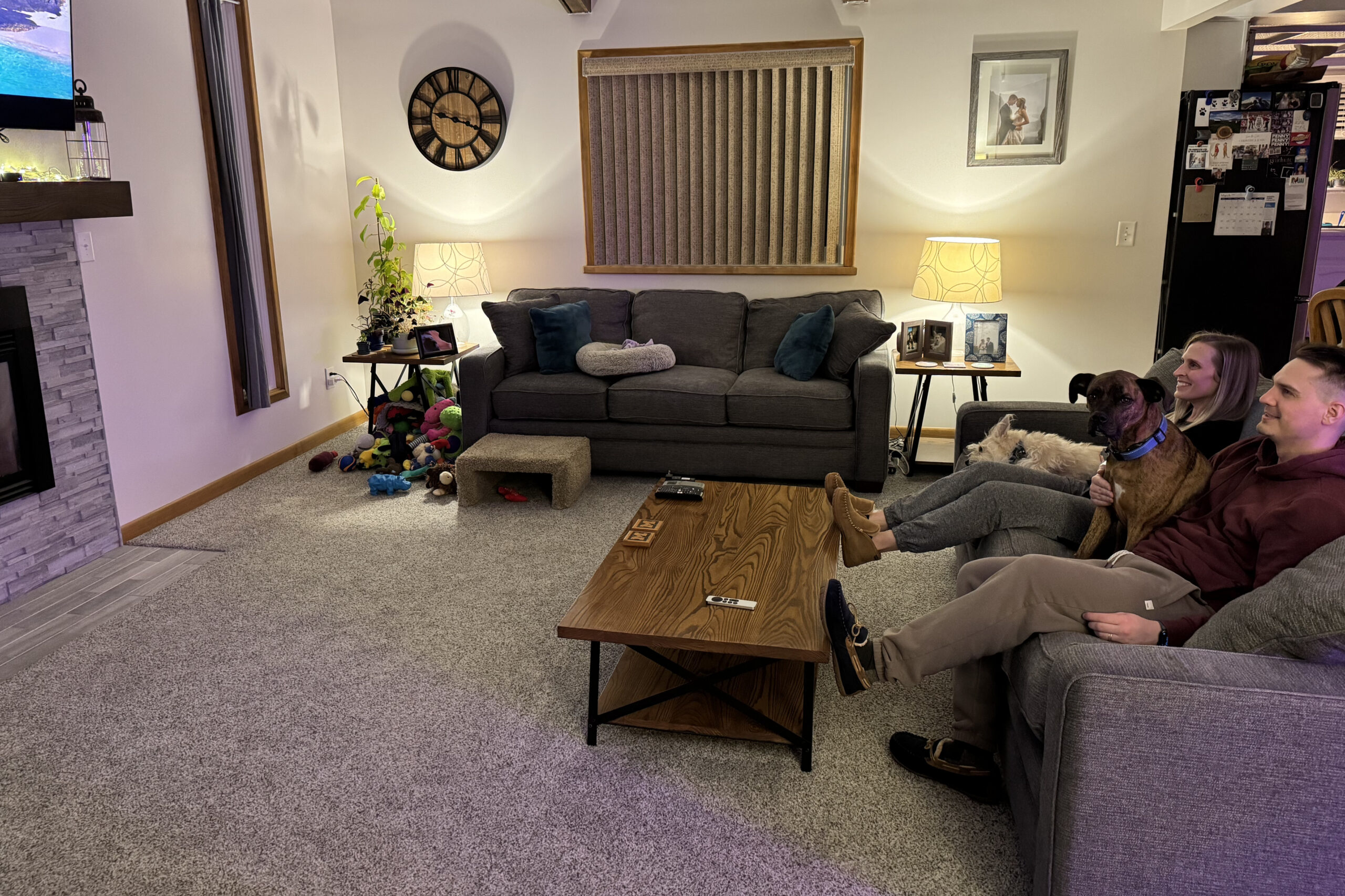 Cozy living room with a gray sectional, two people and two dogs lounging on the right, and a toy-filled carpet nearby.