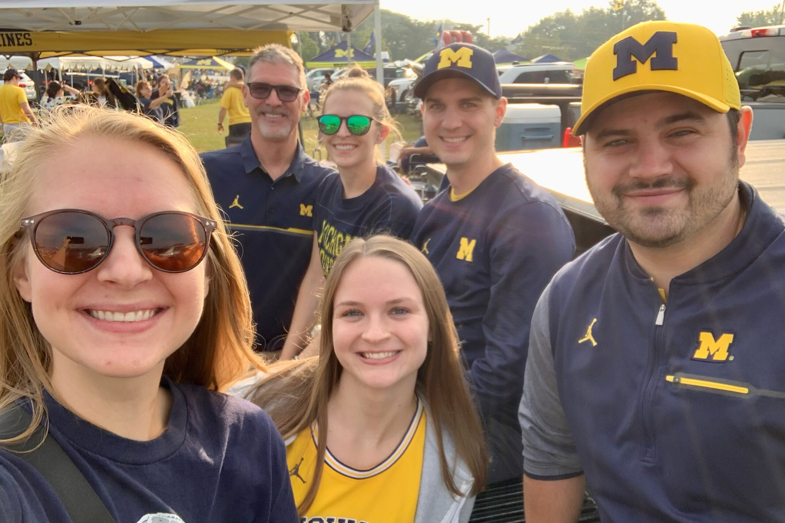 Group selfie at a college event with several people wearing Michigan gear and yellow-blue colors, smiling with tents in the background.