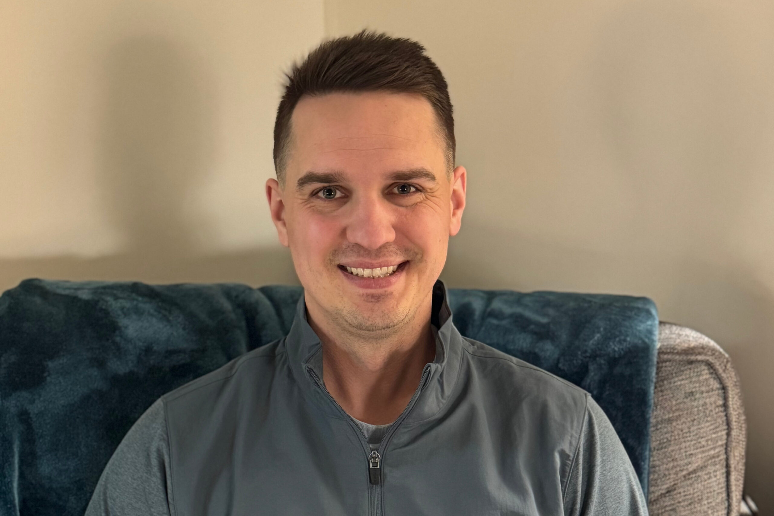 Smiling man taking a selfie on a blue couch with a beige wall behind him.