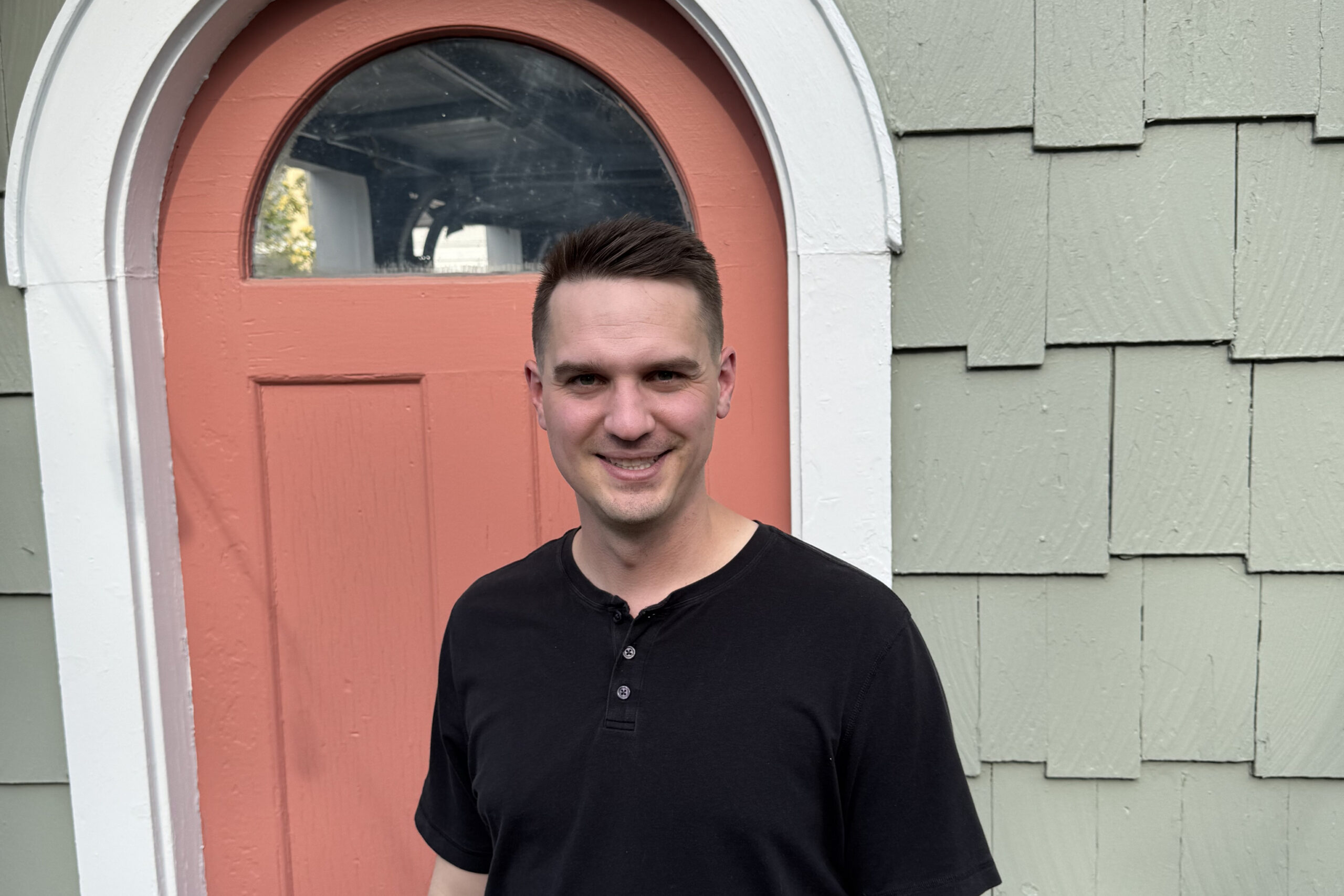 Smiling man in a black shirt standing in front of a peach arched door with white trim.