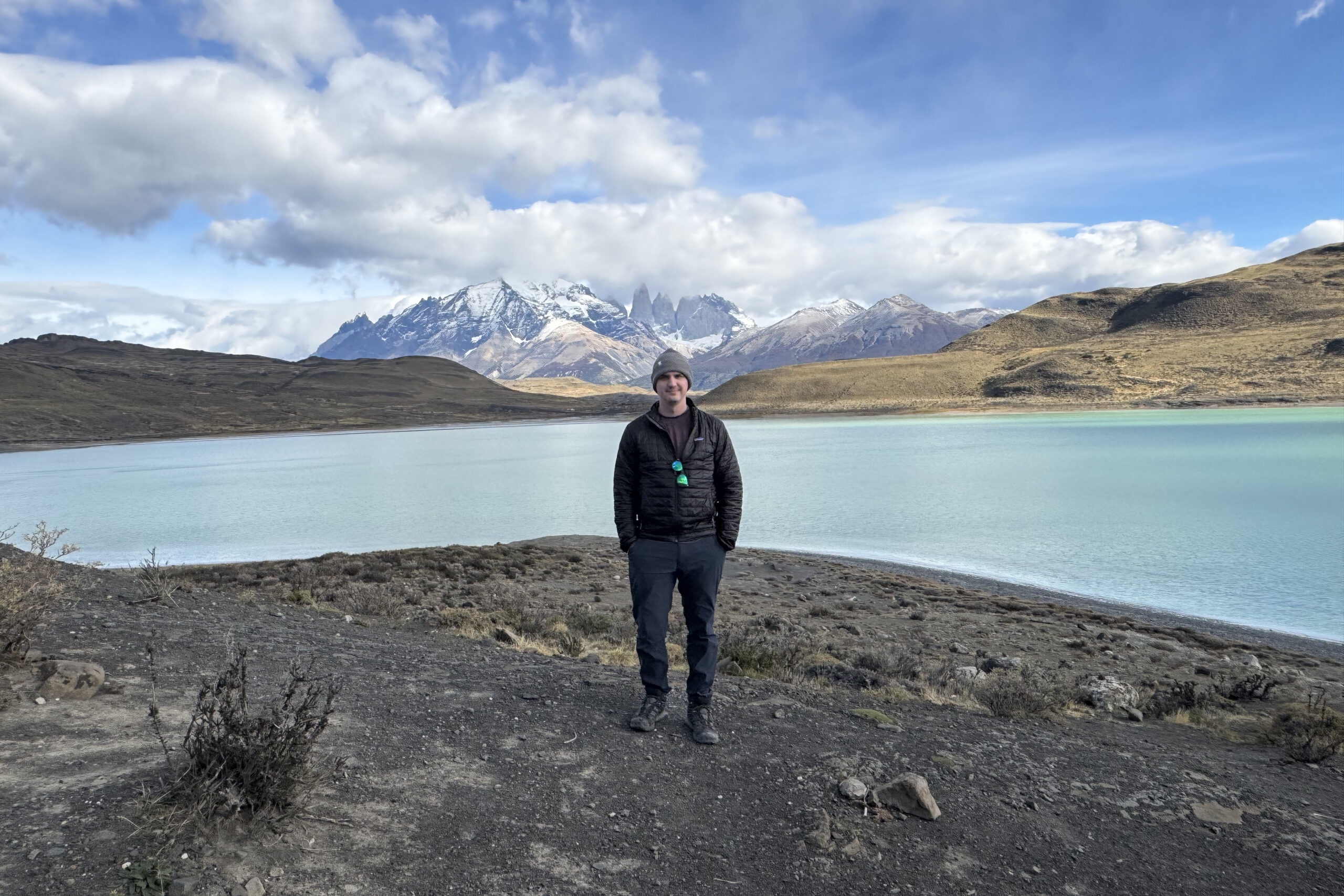 Person standing by a turquoise lake with snow-capped mountains and cloudy sky in the background.