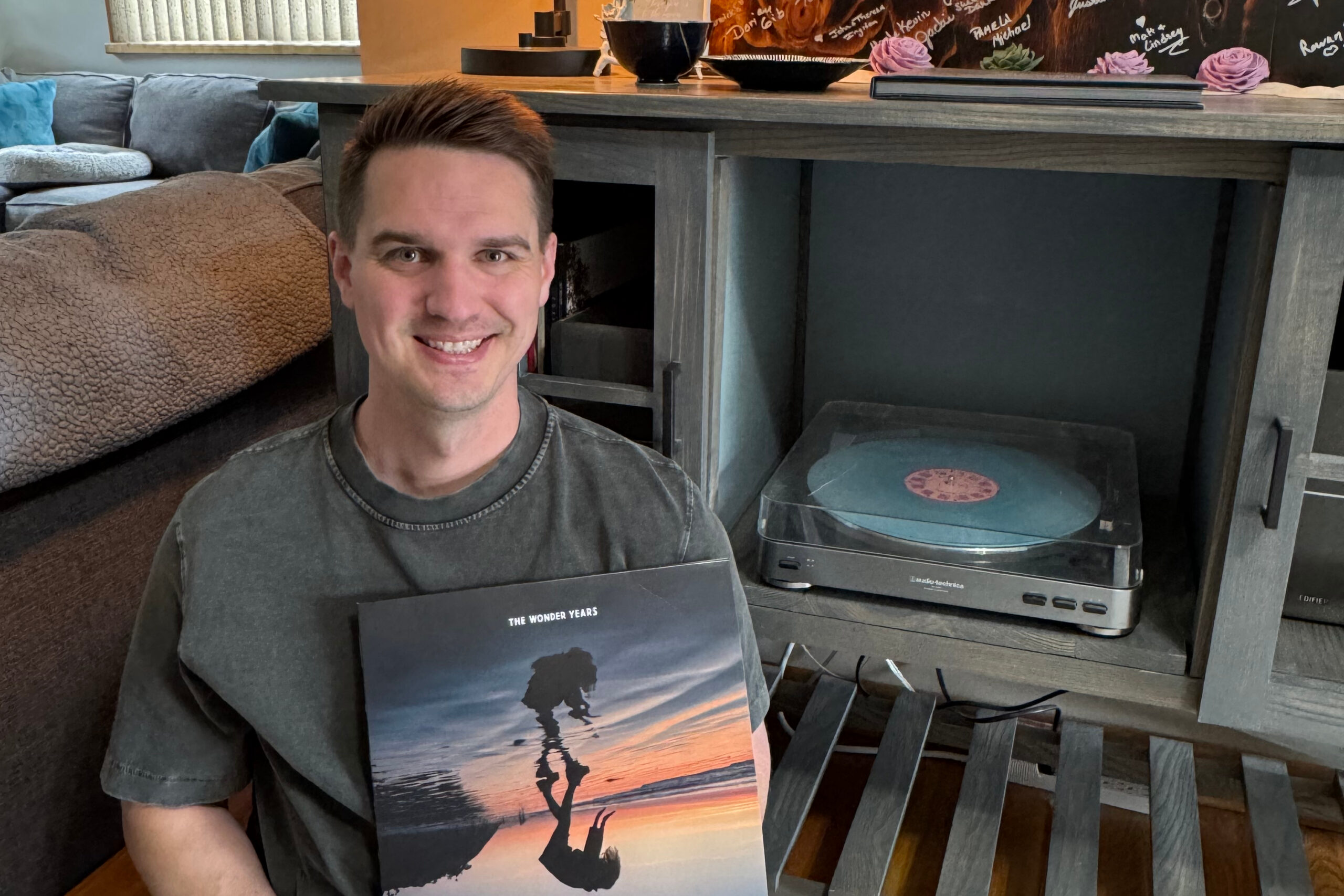 Smiling man in a gray shirt sits indoors, holding a The Wonder Years vinyl with a sunset and jumping silhouette.