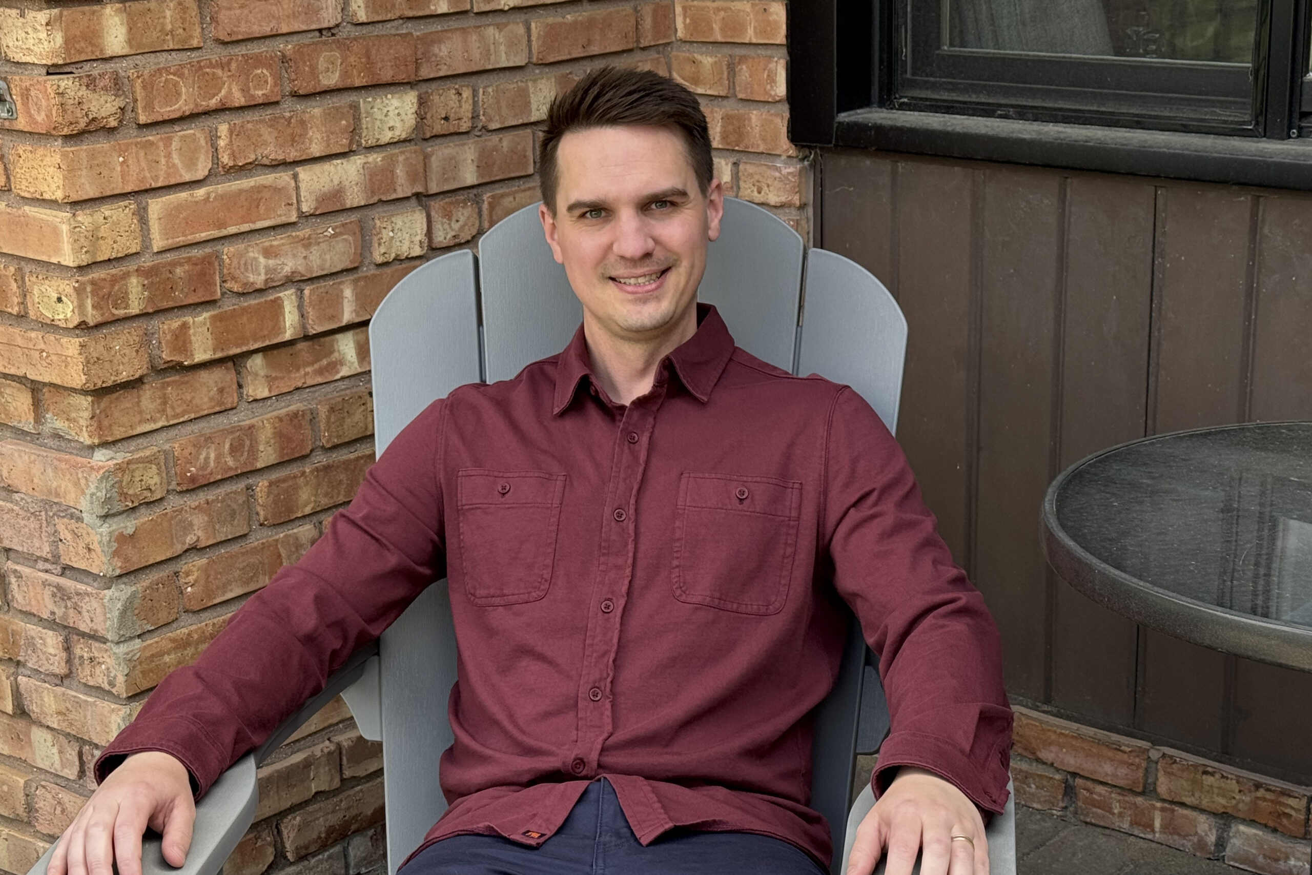 Smiling man in a maroon button-down shirt sits in a gray outdoor chair beside a brick wall and window.
