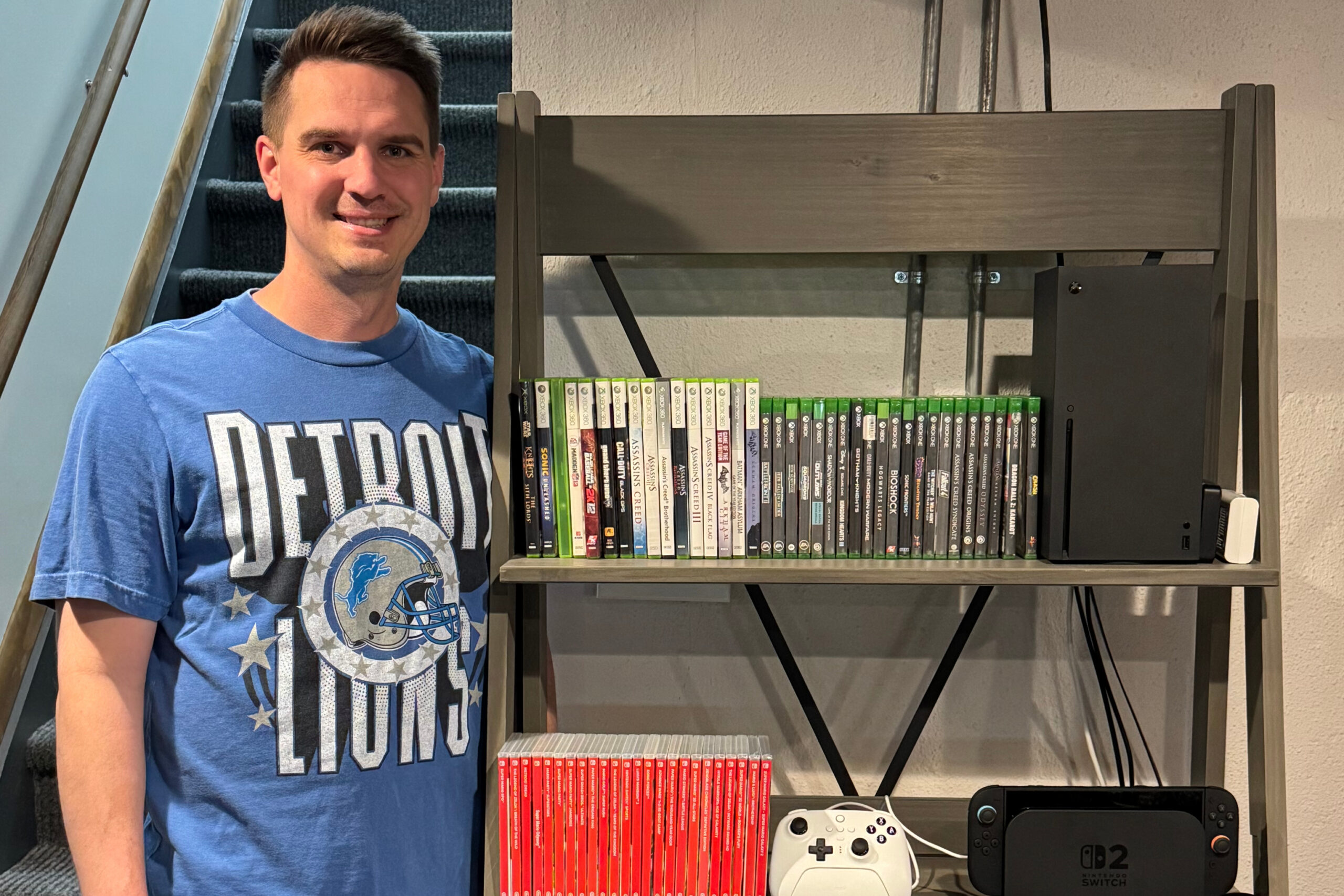 Smiling man in a blue Detroit Lions shirt stands beside a shelf full of video game cases with a white Xbox controller and a Nintendo Switch on the lower shelf.