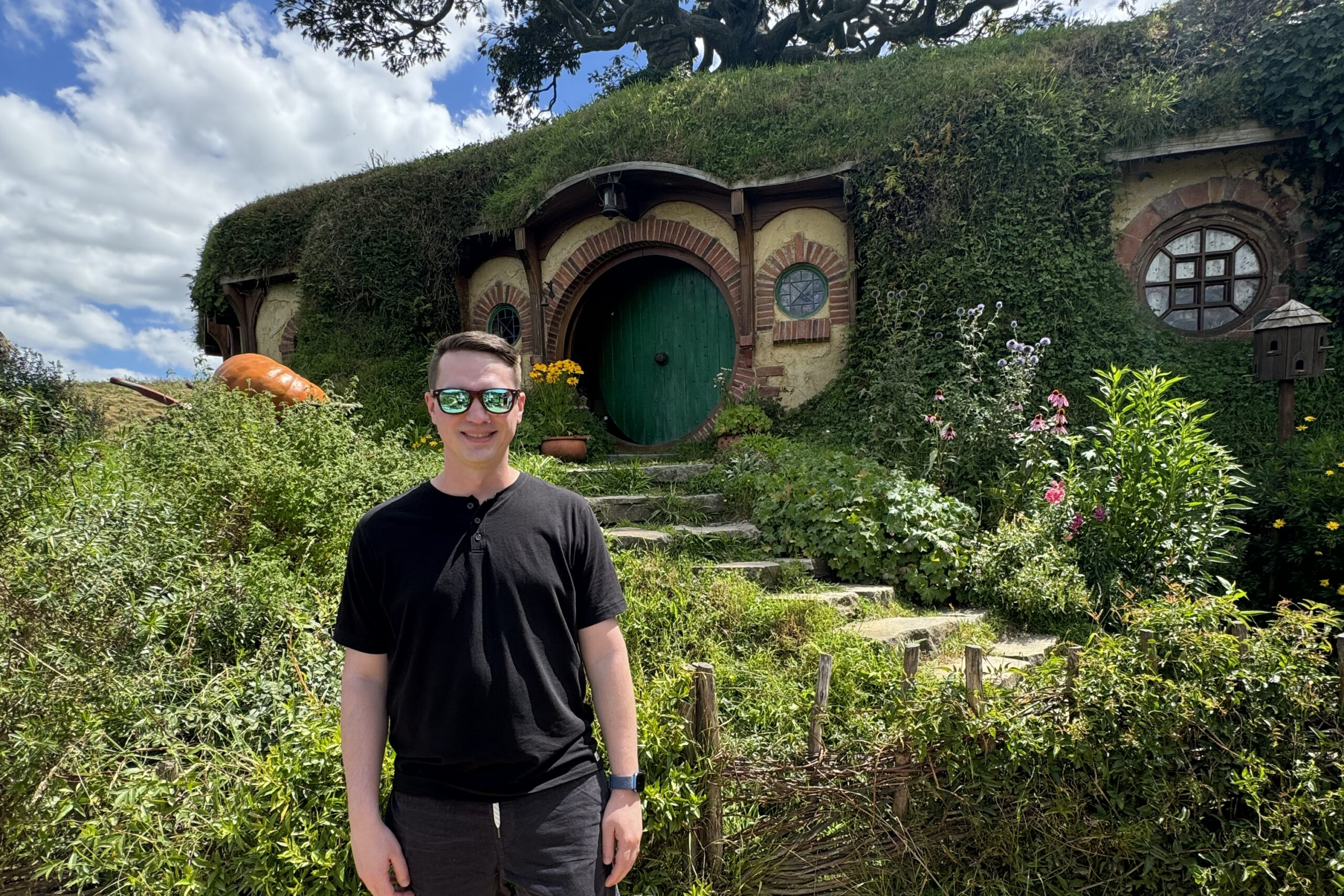 Young man in a black shirt and sunglasses standing in front of a round hobbit-style house with a green door and ivy-covered walls.