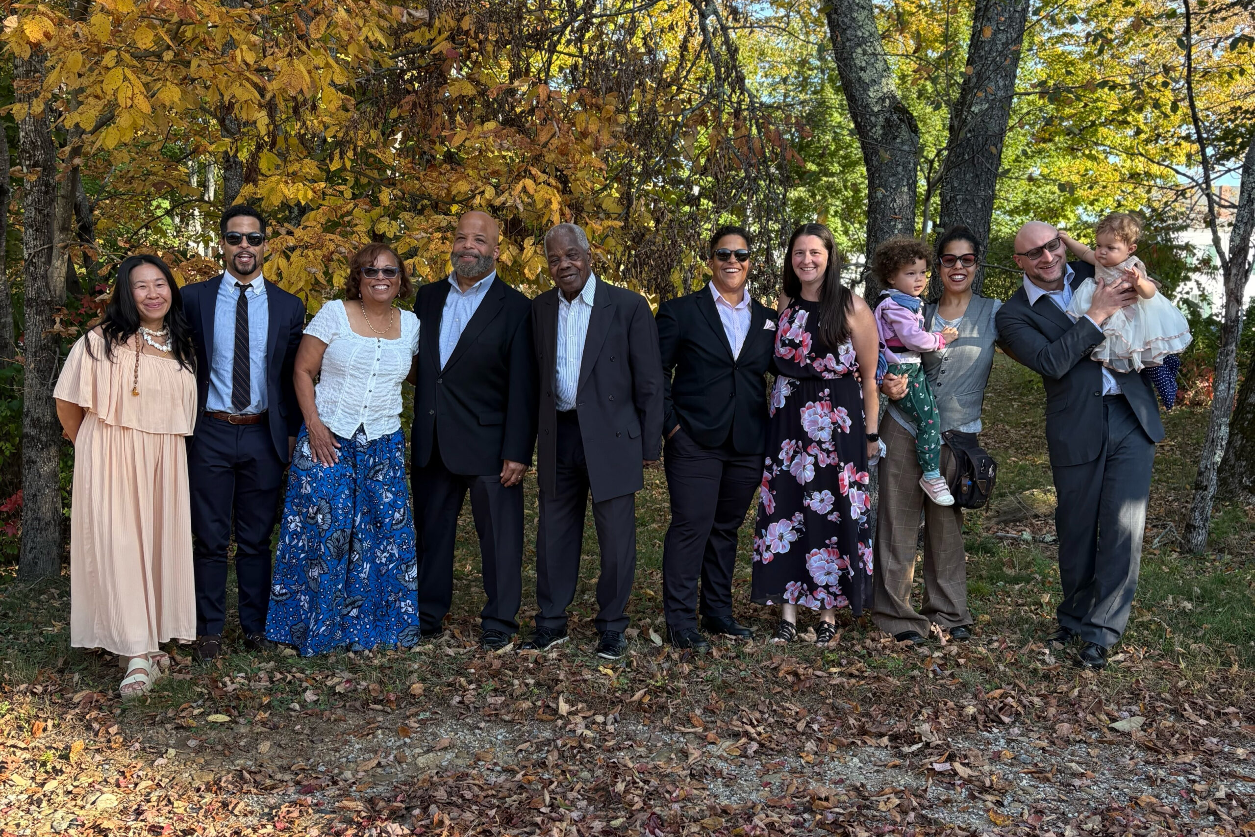 A diverse group of adults and children posing outdoors among autumn leaves in a sunny park.