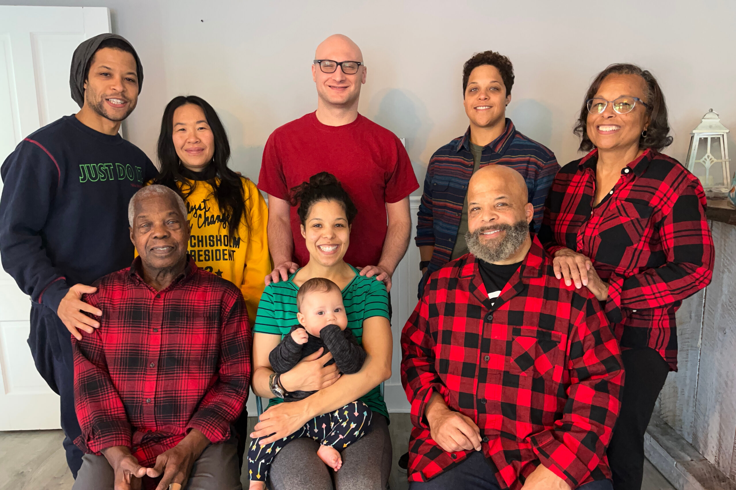 Group of eight people posing together indoors, several wearing red-and-black plaid shirts, a woman holds a baby in a green striped shirt.