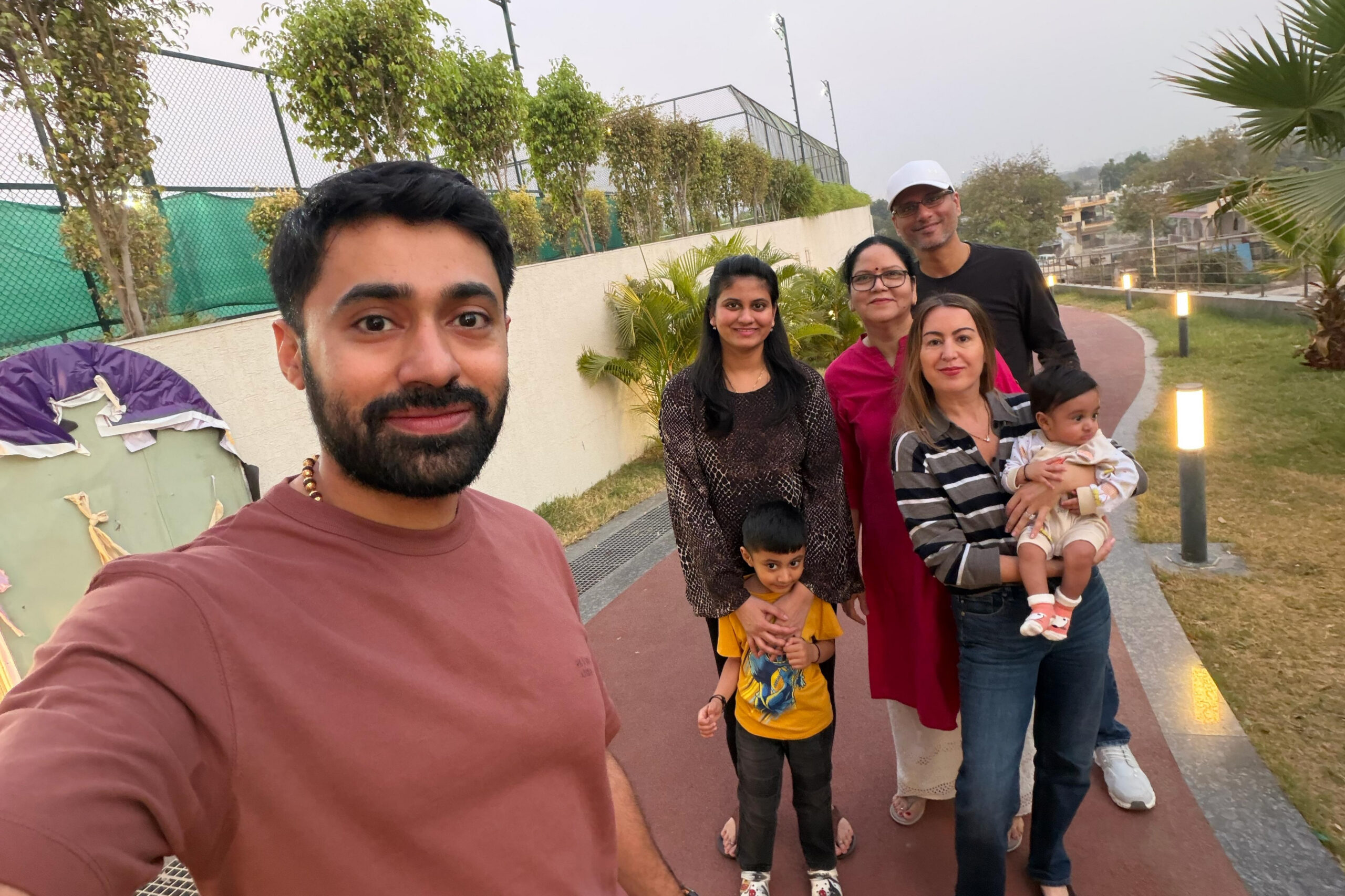 Man in a maroon shirt takes a selfie with a group of six others on a park path; a woman holds a baby while two children stand nearby.