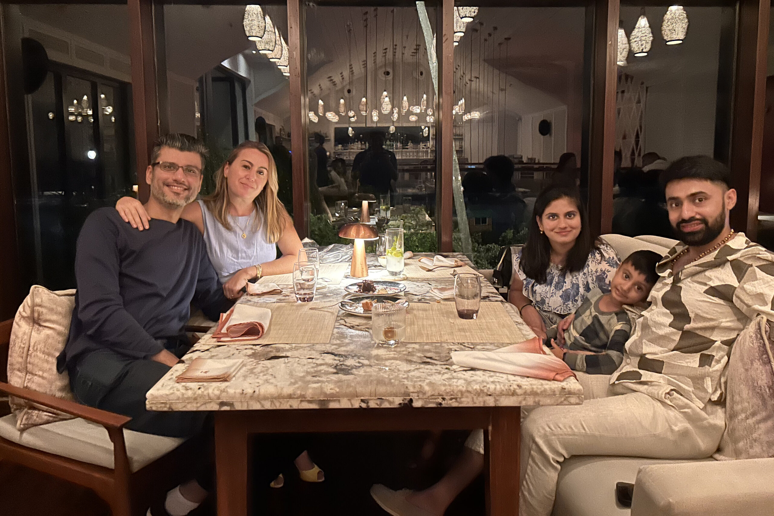 A family of five dining together at a restaurant table with drinks and dessert, smiling at the camera.