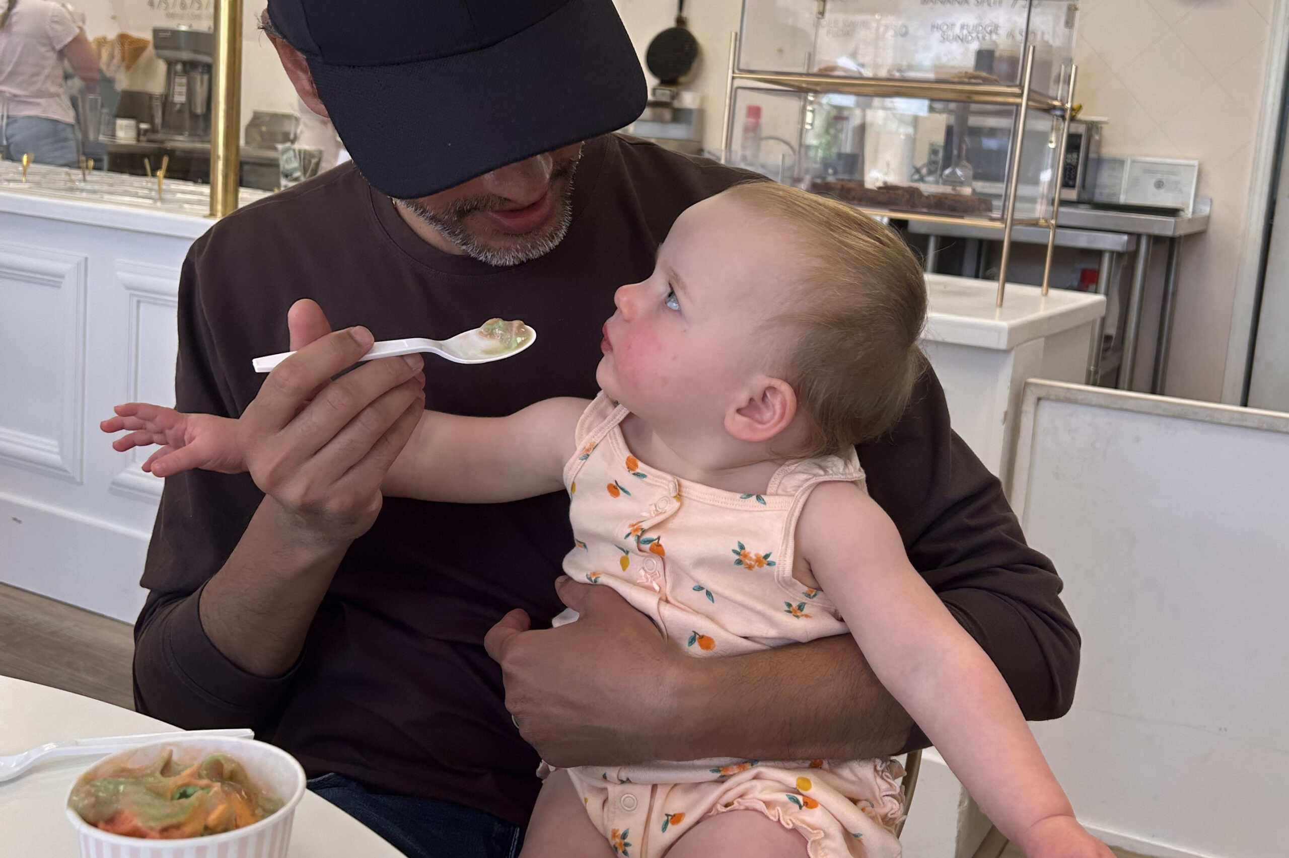Man in a dark cap feeding a baby with a spoon at a cafe; dessert cup on table in foreground.