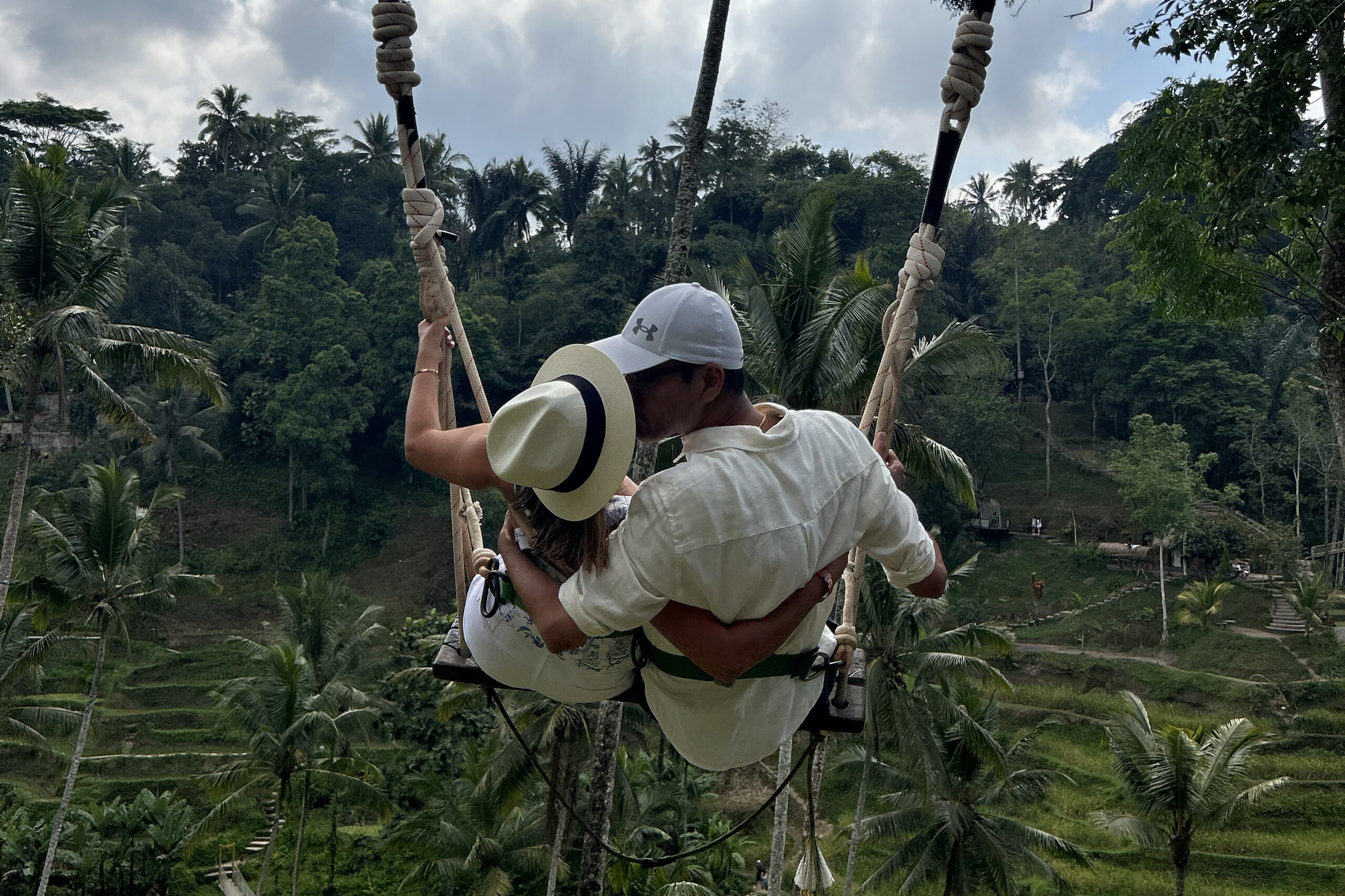 Couple hugging on a rope swing suspended above a tropical hillside with palm trees nearby.
