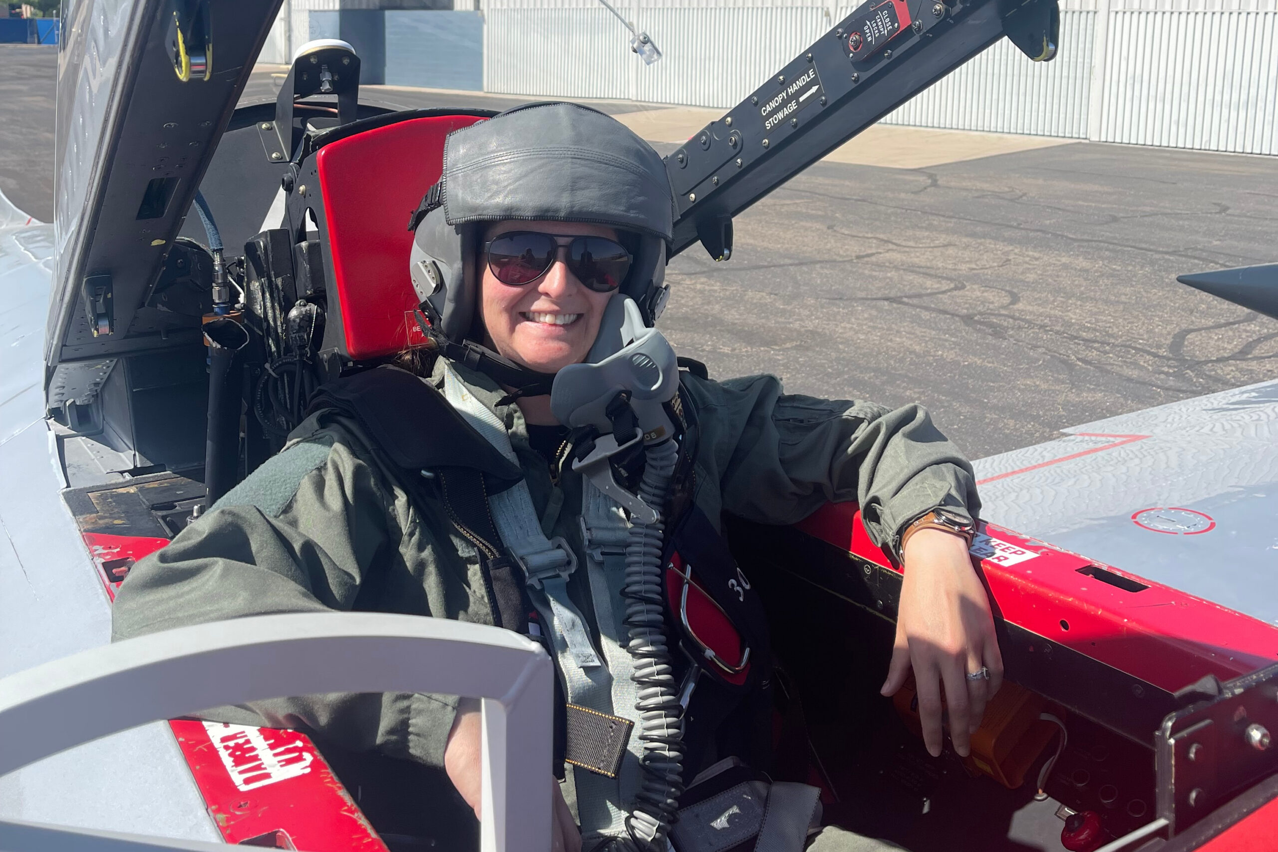 Smiling pilot wearing a helmet and sunglasses sits in the cockpit of a small red airplane on the tarmac.