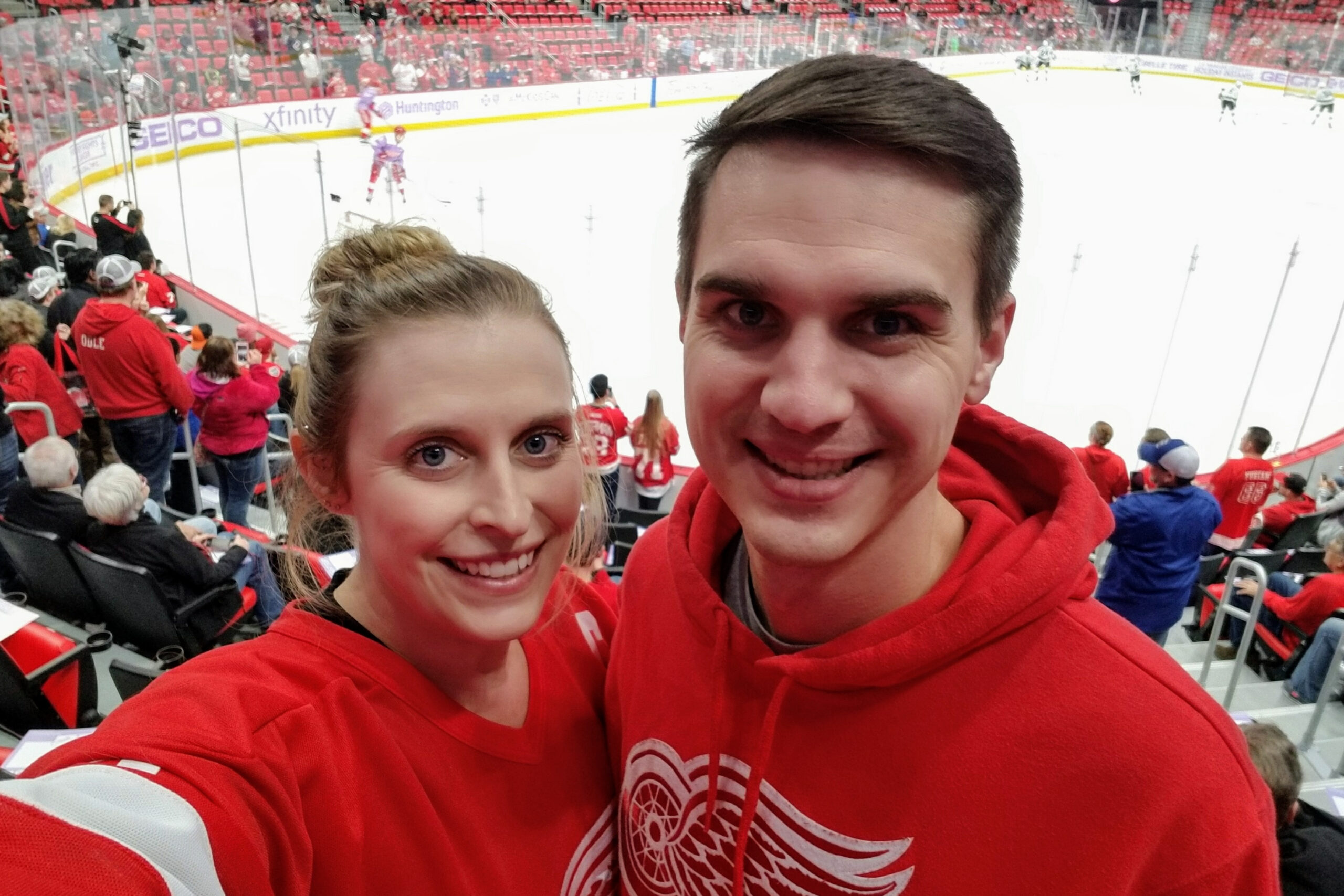 Two fans in red jerseys take a close-up selfie at a hockey game with the ice rink and crowd in red seats behind them.