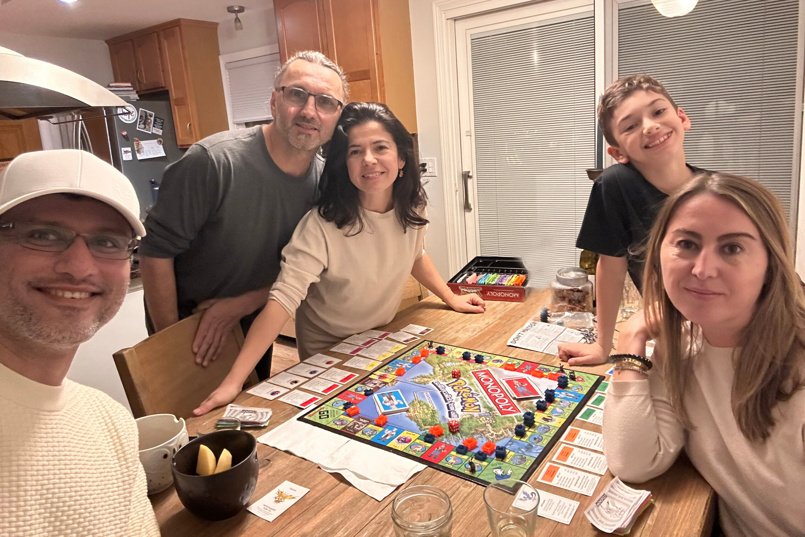 Group of five people smiling around a Monopoly board on a kitchen table, posing for a photo outside the frame of the camera.