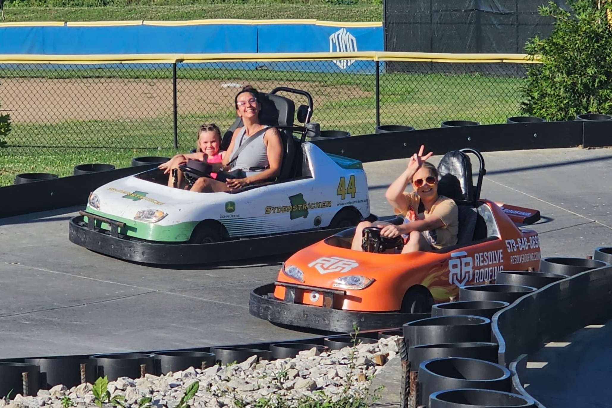 Group of people in go-karts on a race track, smiling and wearing seat belts with a yellow barrier in the background.