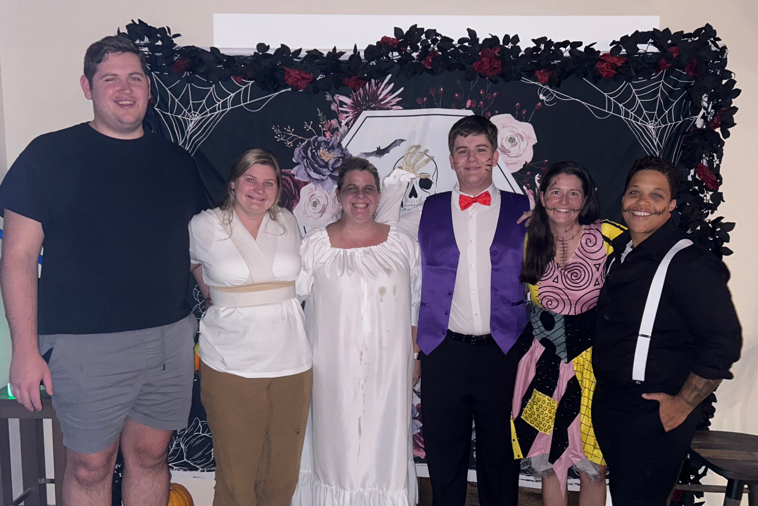 Six adults in Halloween costumes posing together indoors in front of a spiderweb Halloween backdrop with flowers and skull image.