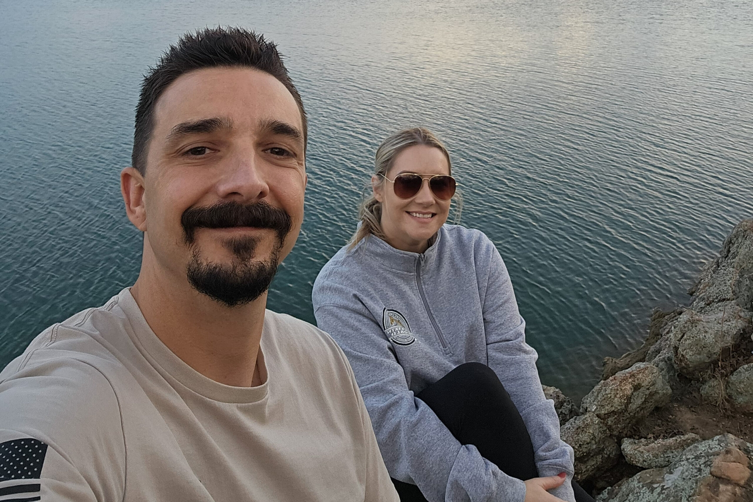 Two people pose for a selfie on rocky shore above calm blue water, wearing casual clothes and sunglasses.