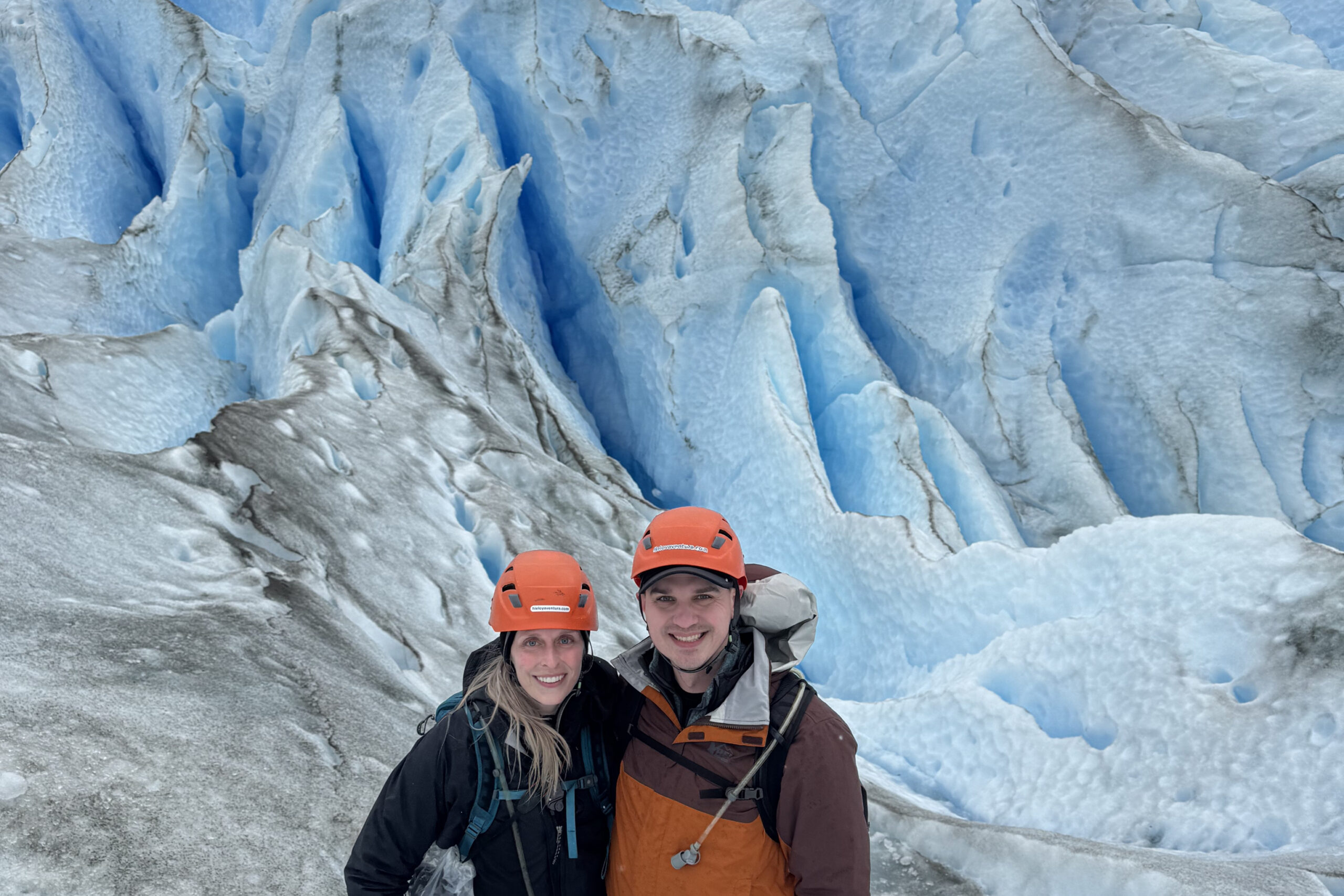 Three hikers wearing orange helmets pose on a blue glacier with jagged ice walls behind them.