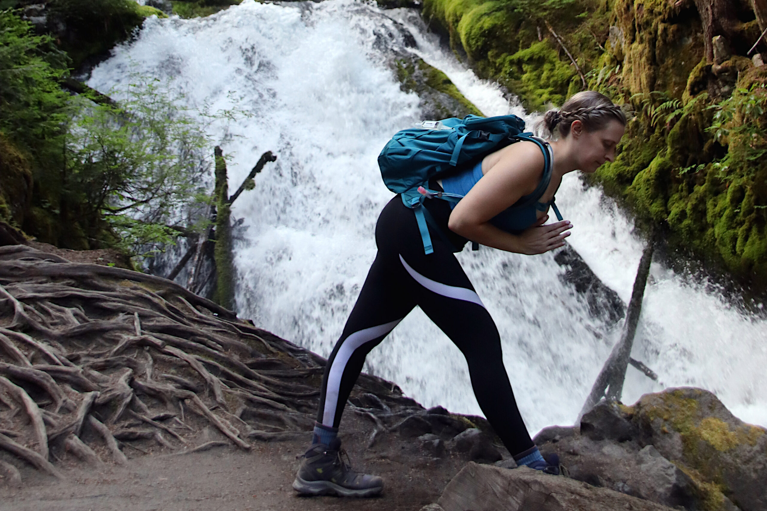 Hiker in a blue backpack bends forward on a rocky trail beside a waterfall.