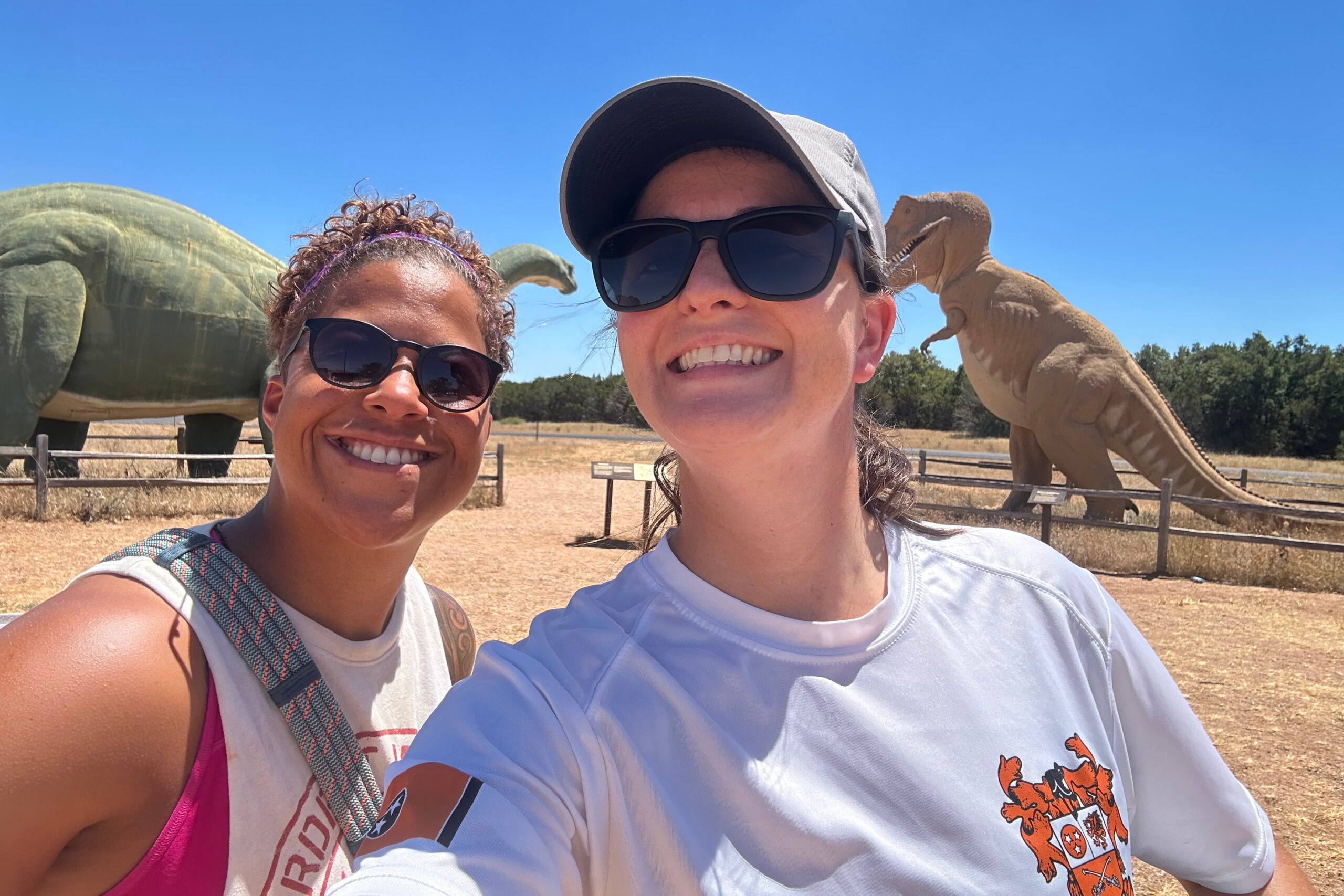 Two people smiling in a sunny outdoor exhibit with large dinosaur statues behind them. blindfolded? No—wearing sunglasses. (Selfie in front of a green dinosaur)