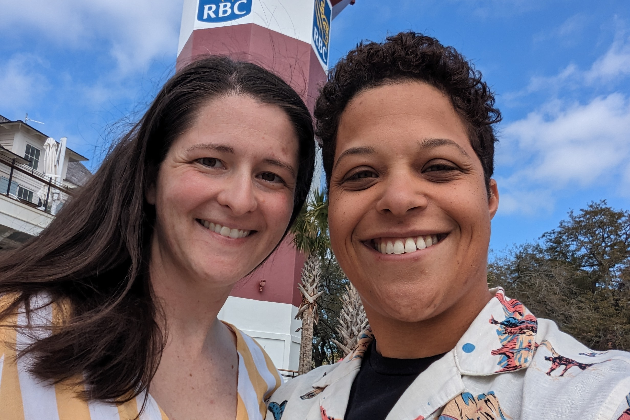 Two people smiling for a selfie outdoors with a blue sky and an RBC sign on a red-and-white building behind them.