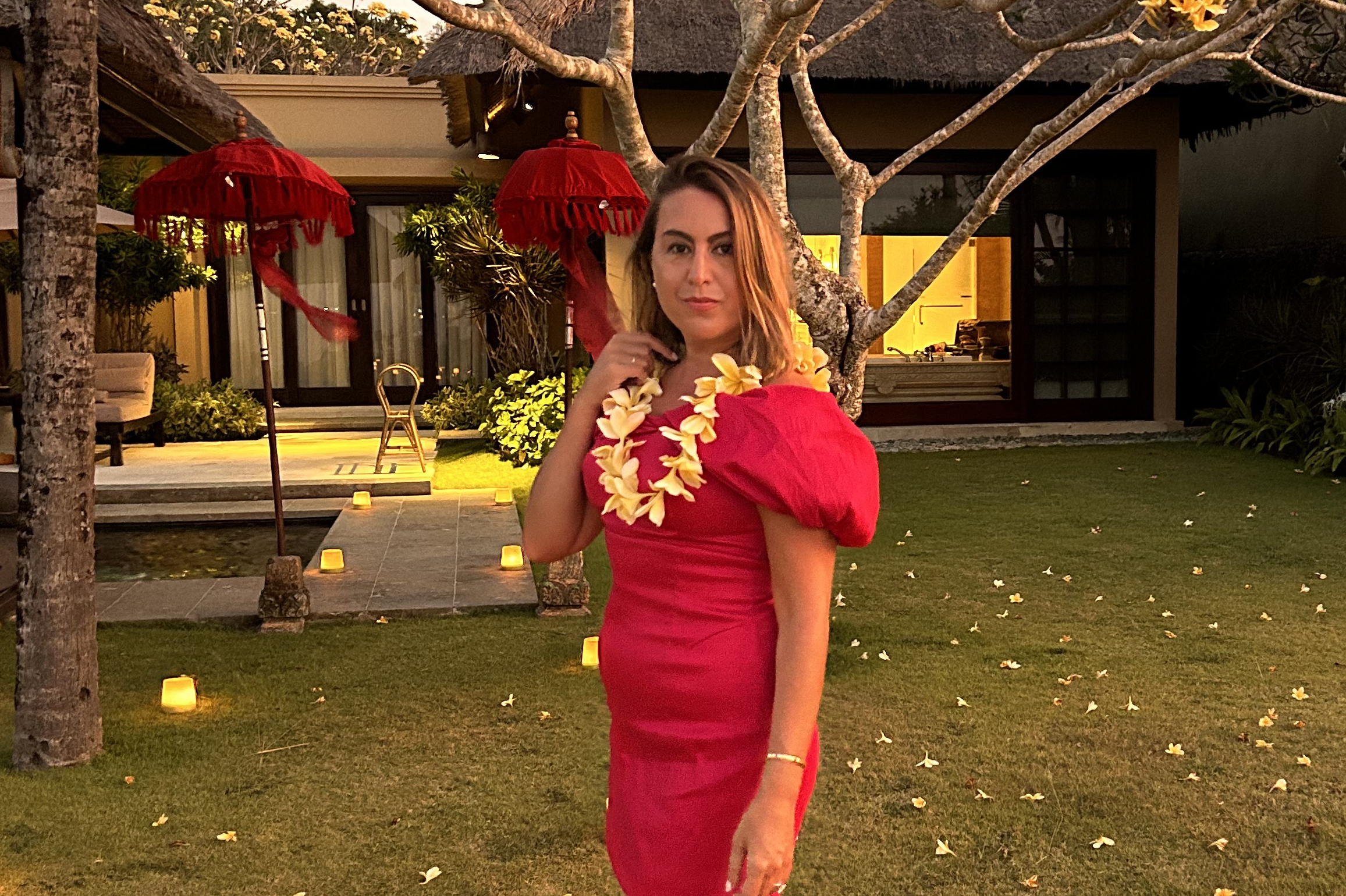 Woman in a red dress wearing a yellow flower lei, standing in a tropical garden with tiki torches and a thatched building in the background.