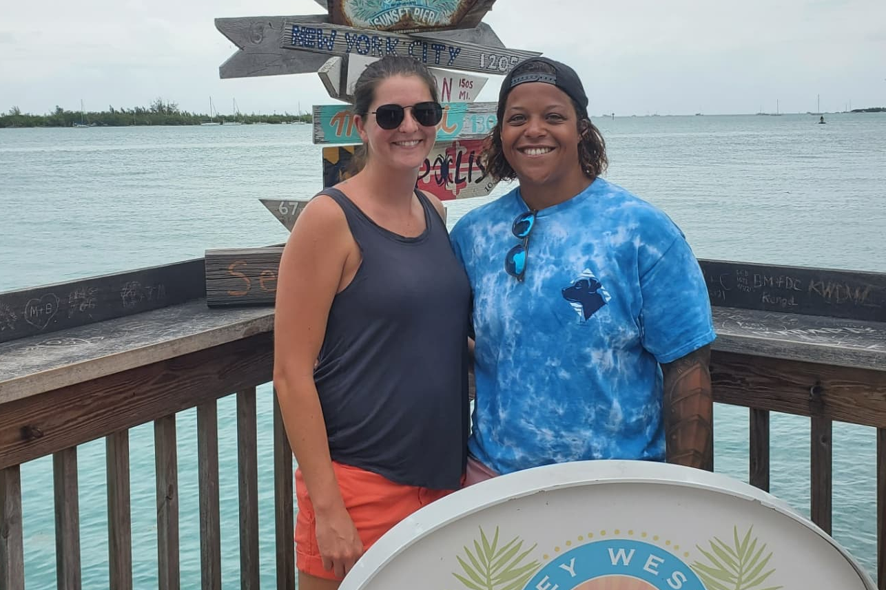 Two people stand on a marina deck by turquoise water, smiling, with a colorful signpost in the background.