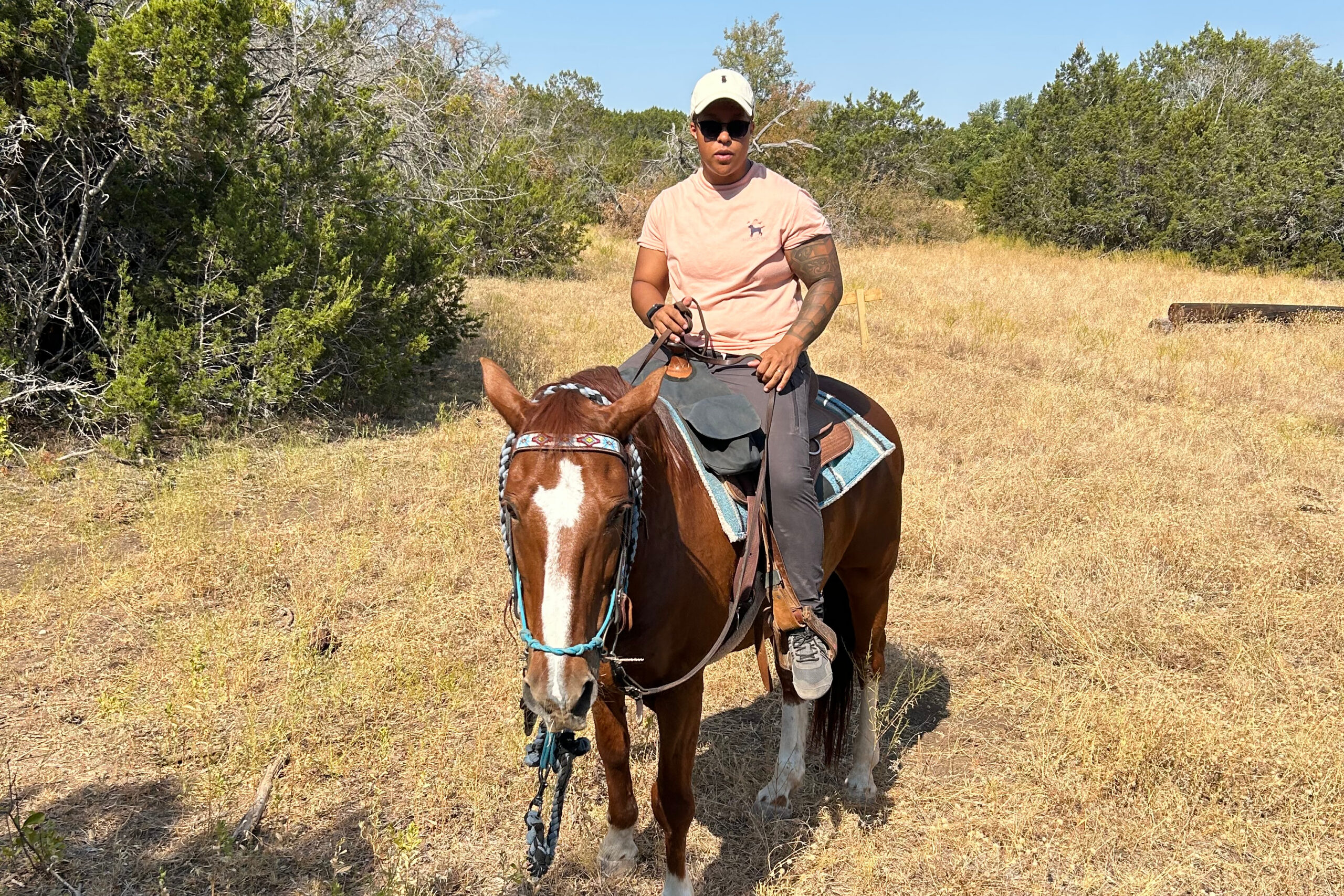 Man wearing a peach t-shirt, cap, and sunglasses rides a brown horse through a dry grassy field with trees in the background.