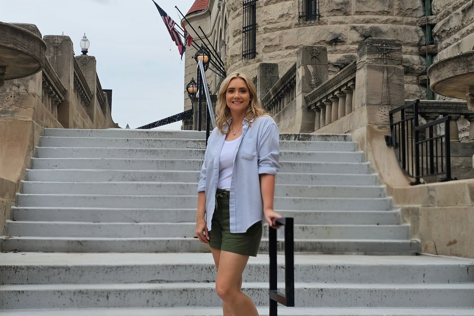 Woman in a light blue shirt over a white top standing on wide stone stairs beside a historic stone building with flags.
