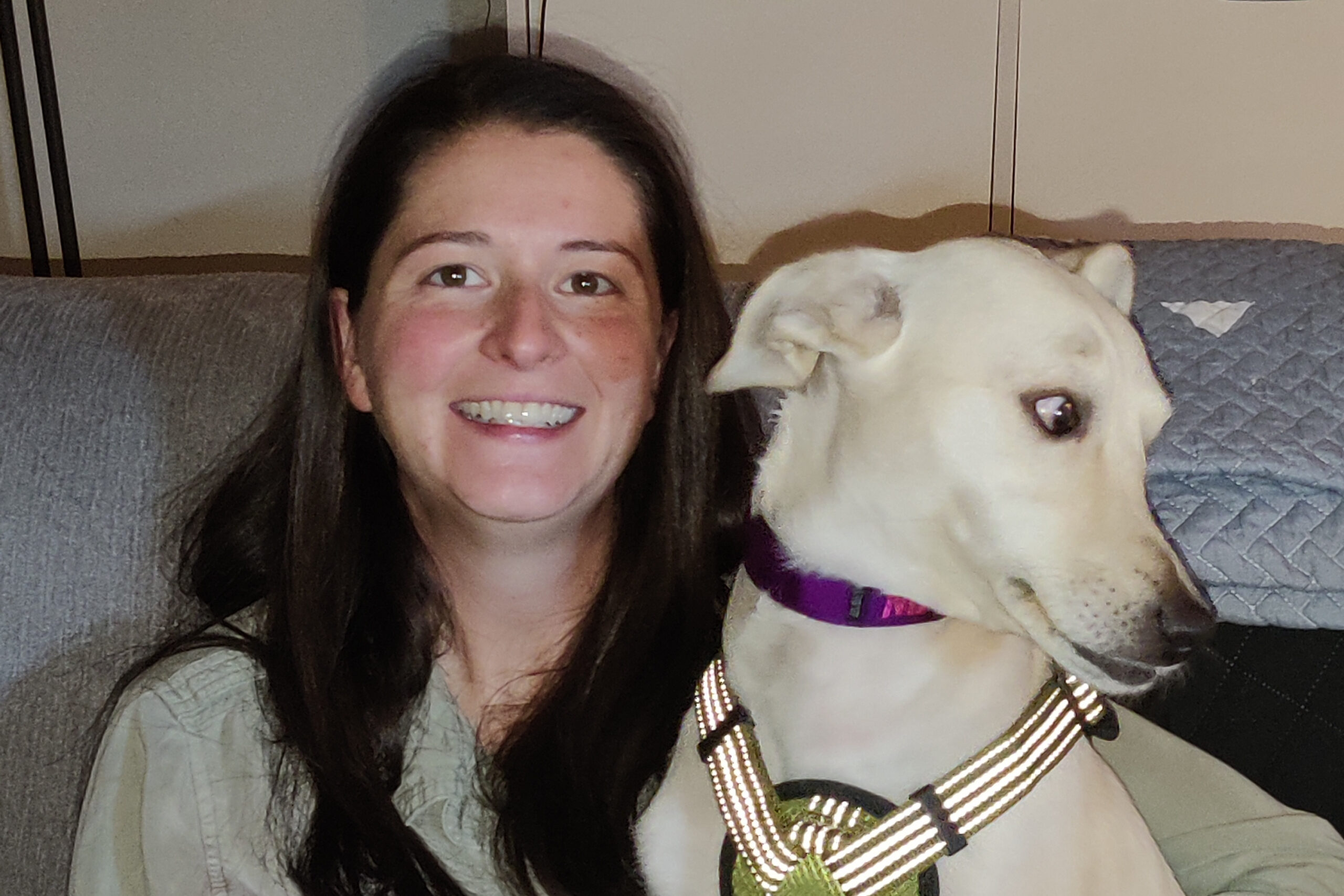Smiling woman on a couch with a white dog wearing a purple collar beside her.