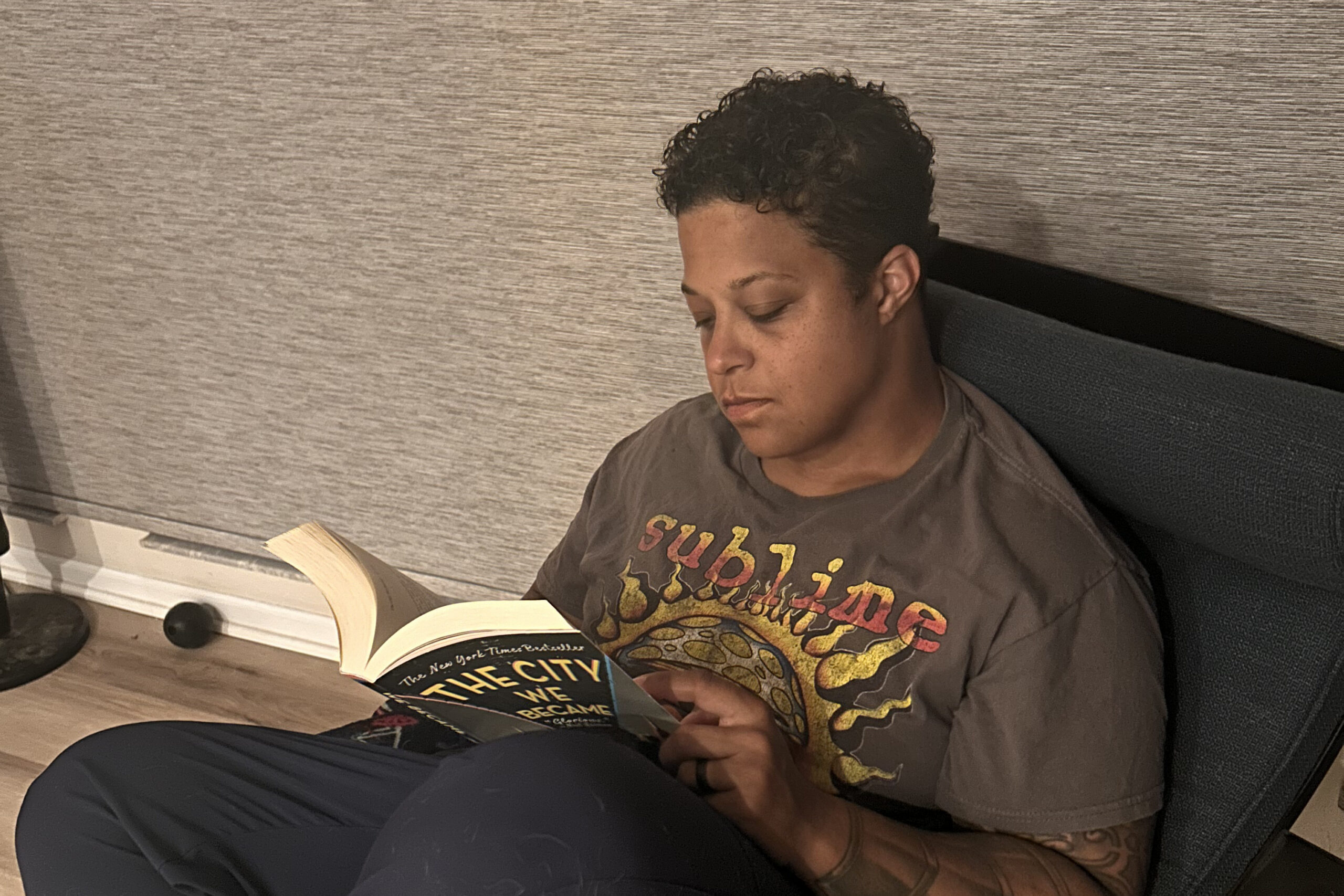 Person sits on a couch reading a paperback, concentrating on a book titled 'The City We Became' with a curved edge cover visible.