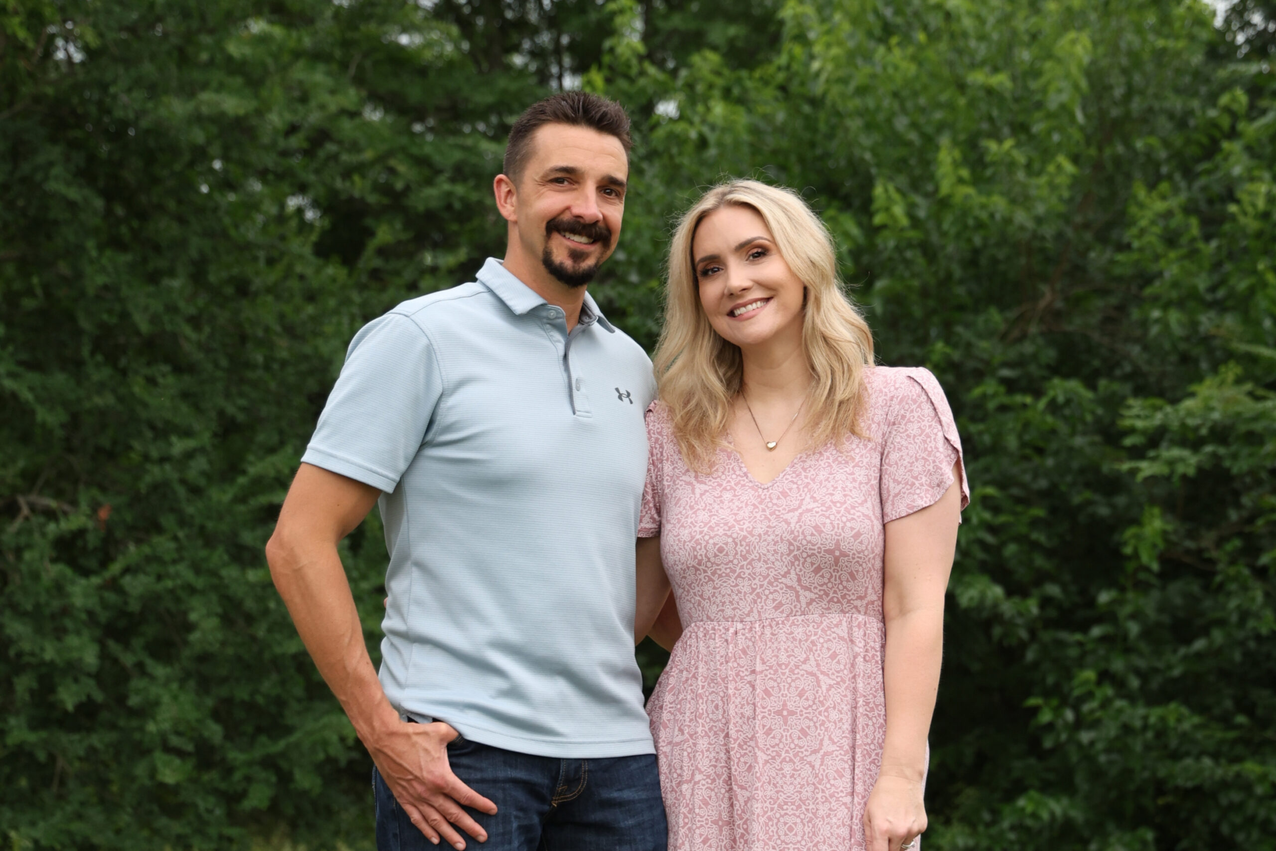 Smiling couple outdoors in front of green bushes; man in light blue polo, woman in pink dress.