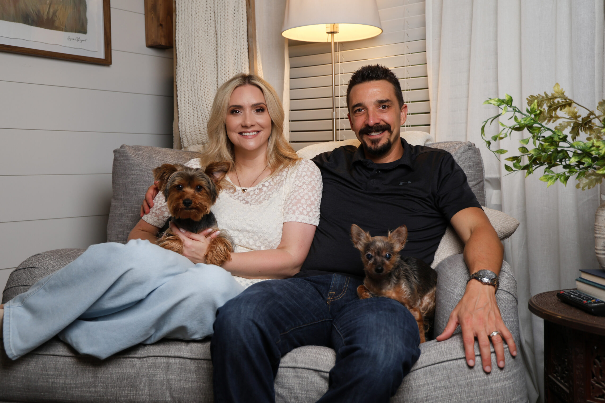 Smiling couple sitting on a gray couch with two small dogs in their laps, cozy living room with lamp and plants behind them.