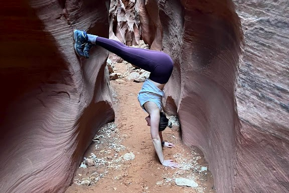 Person in a light blue top and purple leggings holds a handstand between narrow sandstone canyon walls with a sandy floor.
