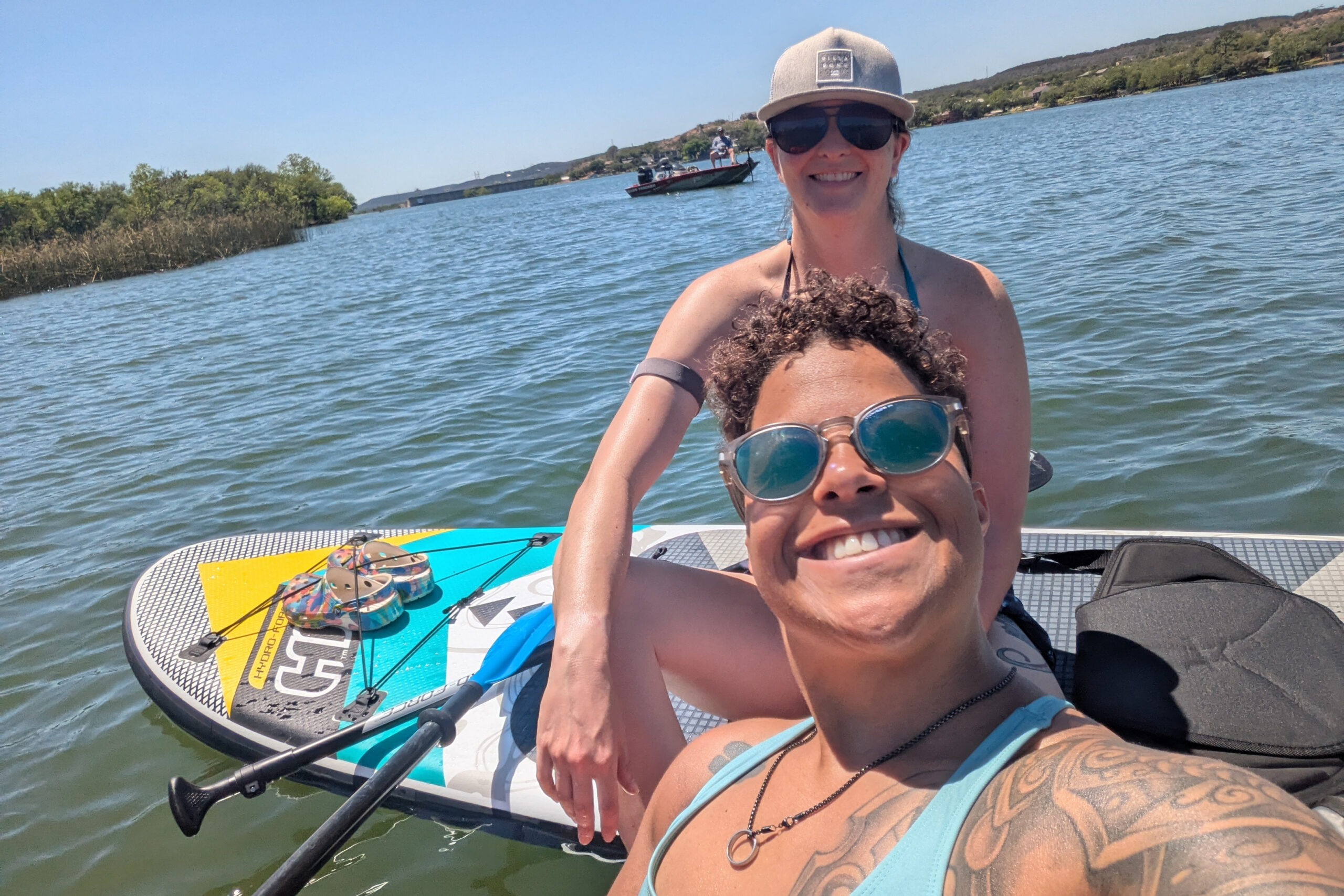 Two friends selfie on a paddleboard over blue water, smiling with sunglasses on a sunny day.