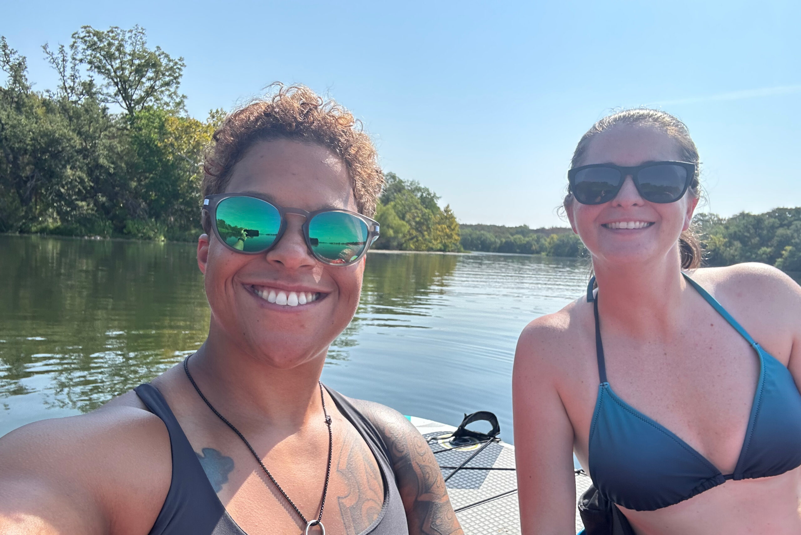 Two women in swimsuits wearing sunglasses taking a selfie on a boat over a calm lake with trees and a blue sky behind them