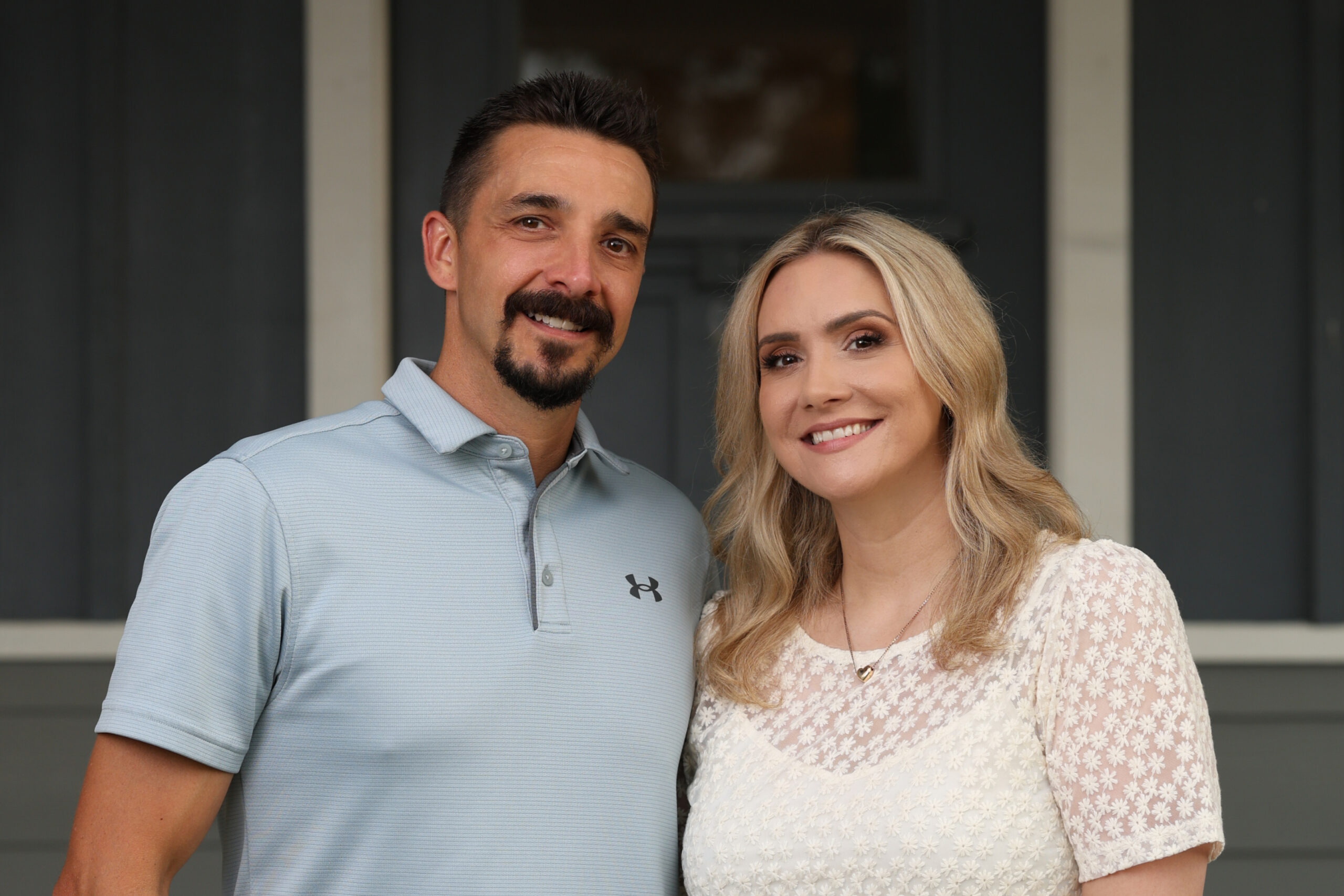Smiling couple posing outdoors in front of a dark gray home facade; man in light blue polo, woman in white lace top with blonde hair