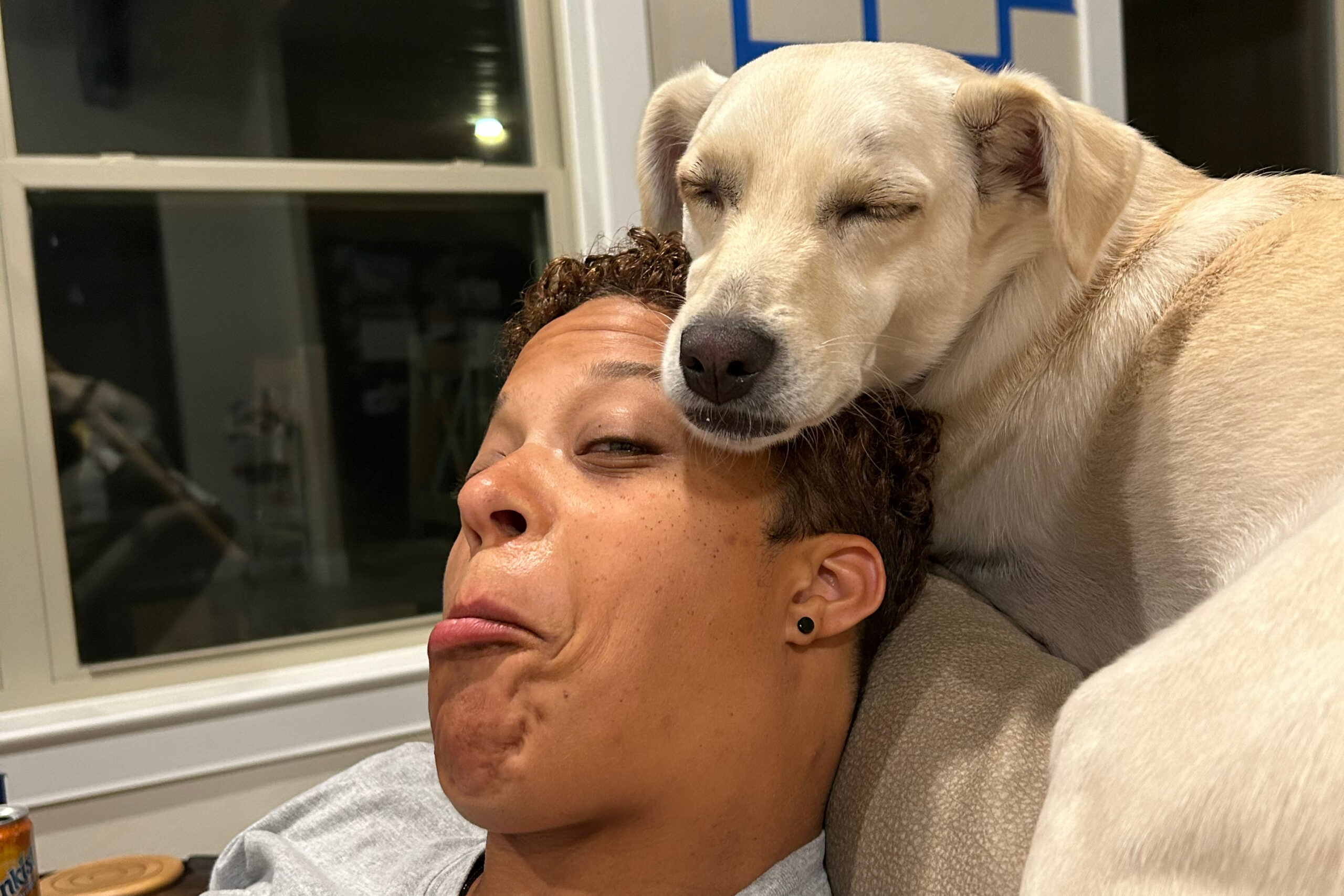 A person with a light-colored dog resting its head on the person's shoulder, both appearing relaxed indoors near a window.