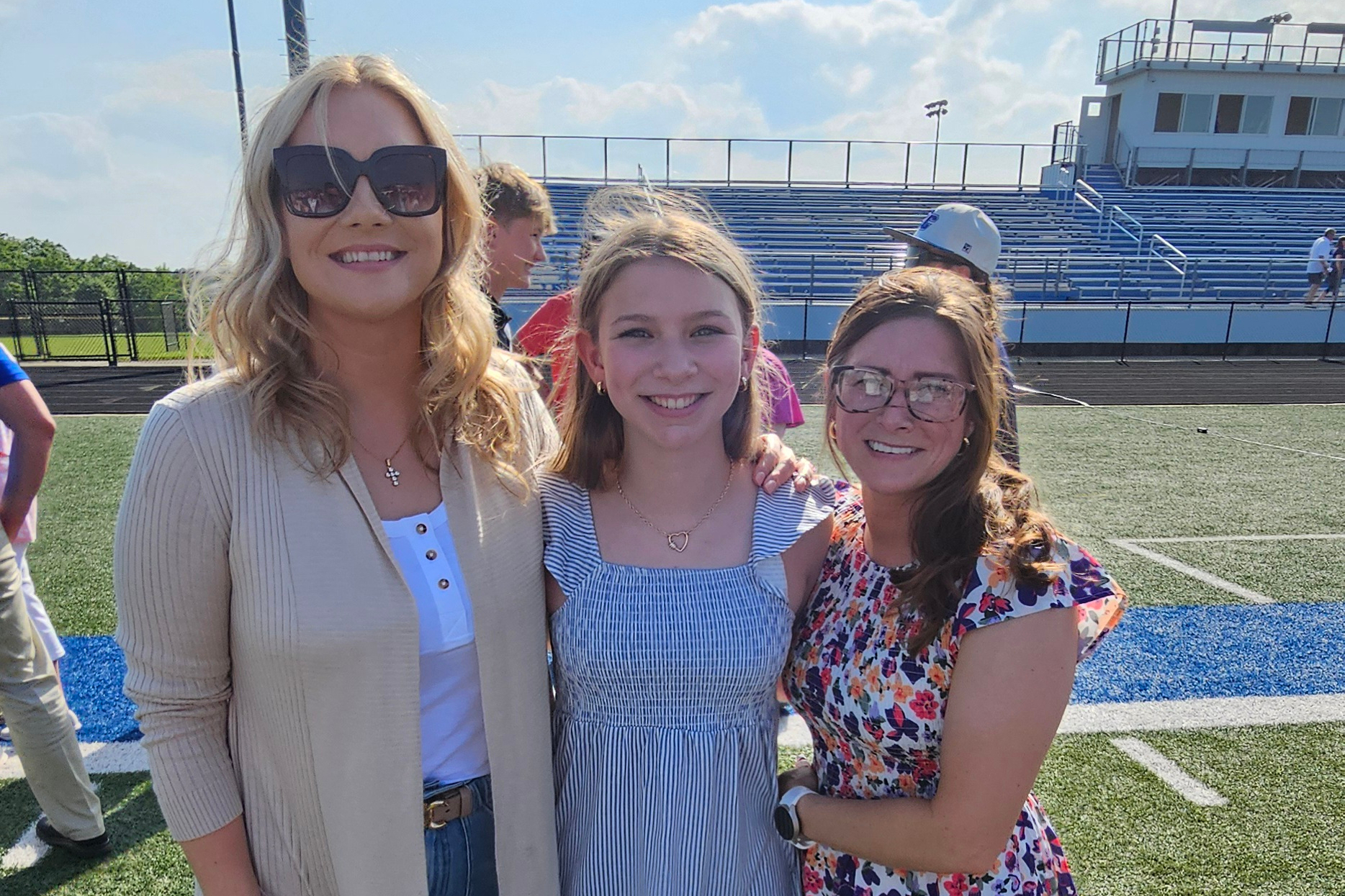 Three smiling friends pose together on a sunny sports field with blue bleachers in the background.