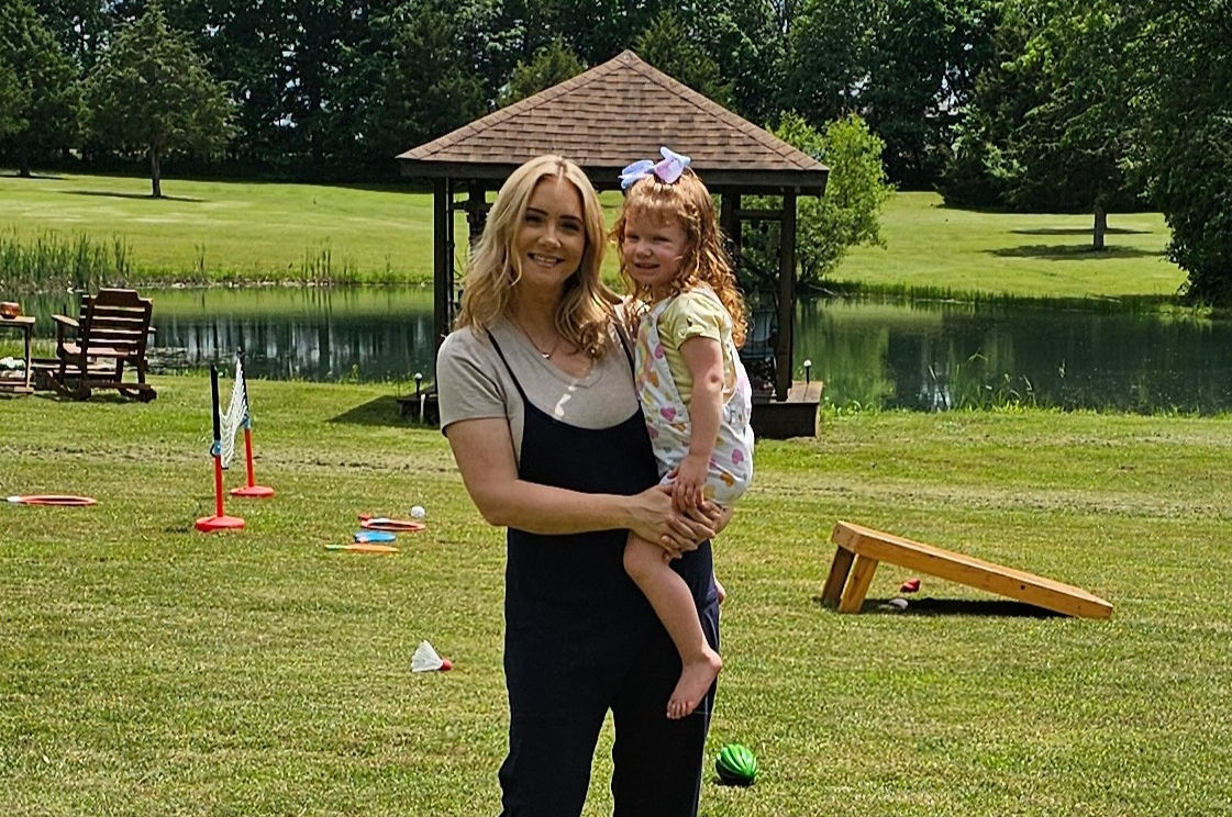 Woman holding a smiling young girl outdoors on a grassy park with a pond and gazebo in the background.