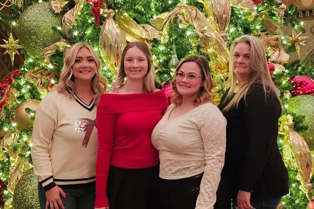 Four women stand together in front of a highly decorated Christmas tree with gold ribbons and lights, smiling for the photo, wearing festive outfits.