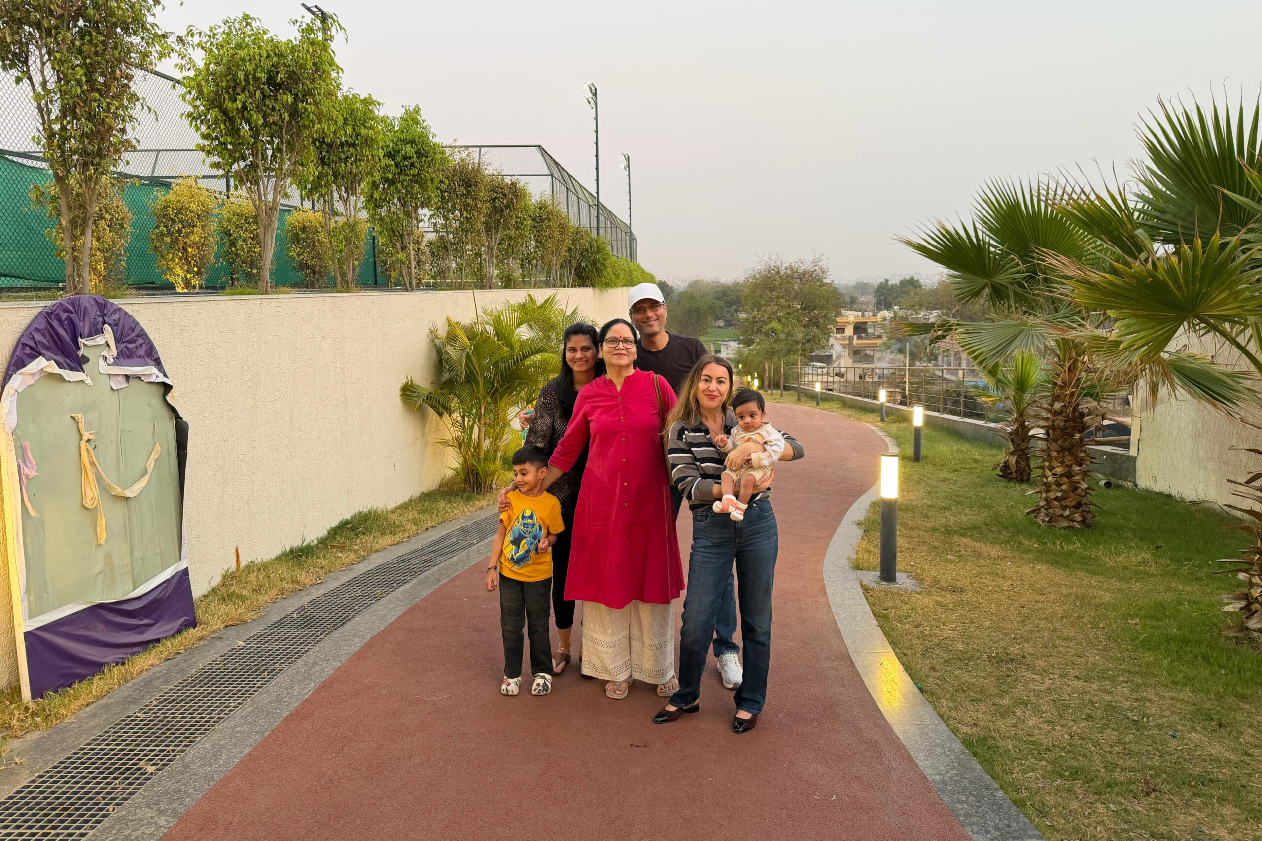 Group of six people posing on a red walking path beside a tan wall and palm trees in a park setting.