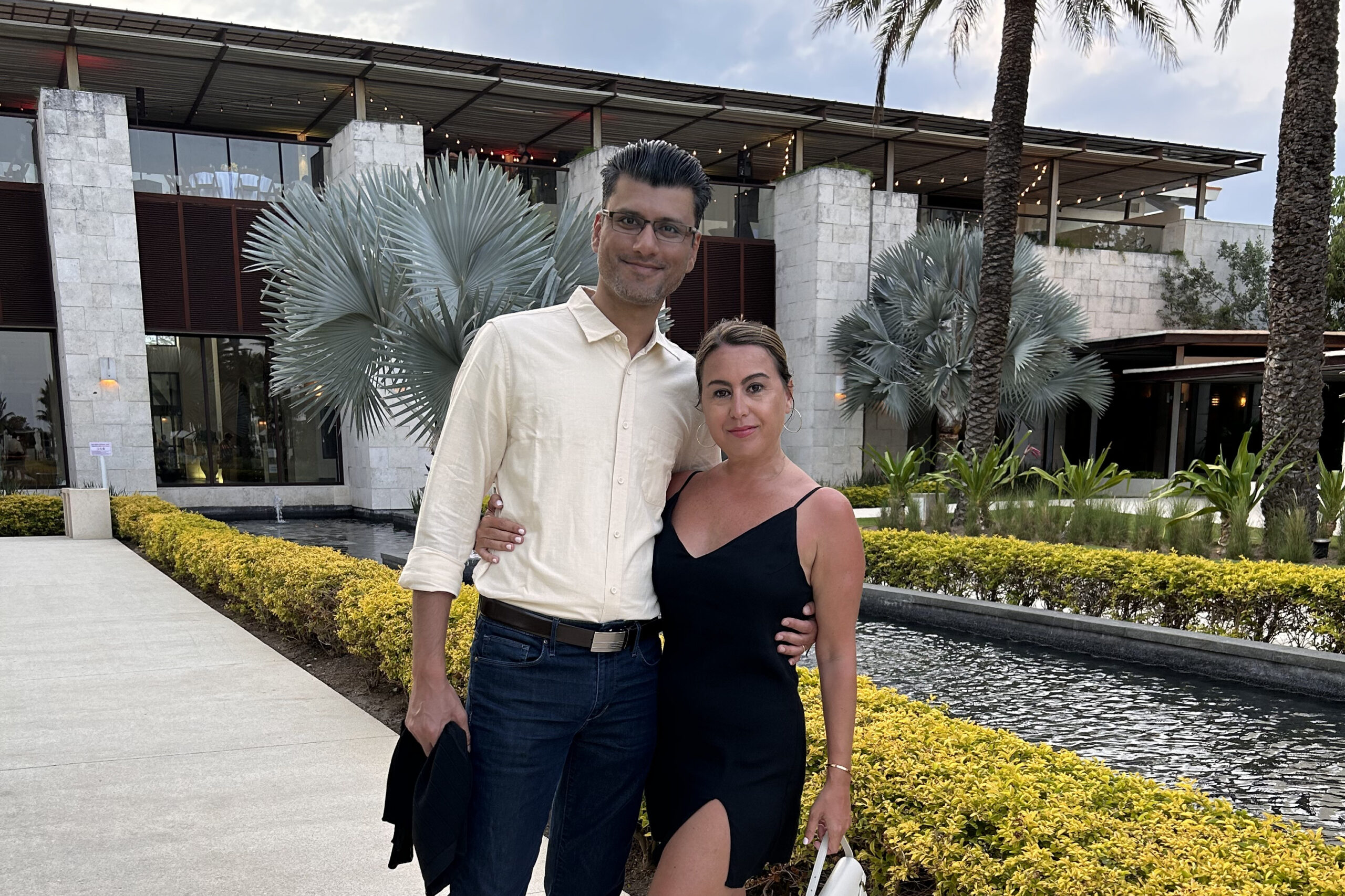 A smiling couple stands together outdoors in front of a modern building with palm trees and yellow shrubs behind them.