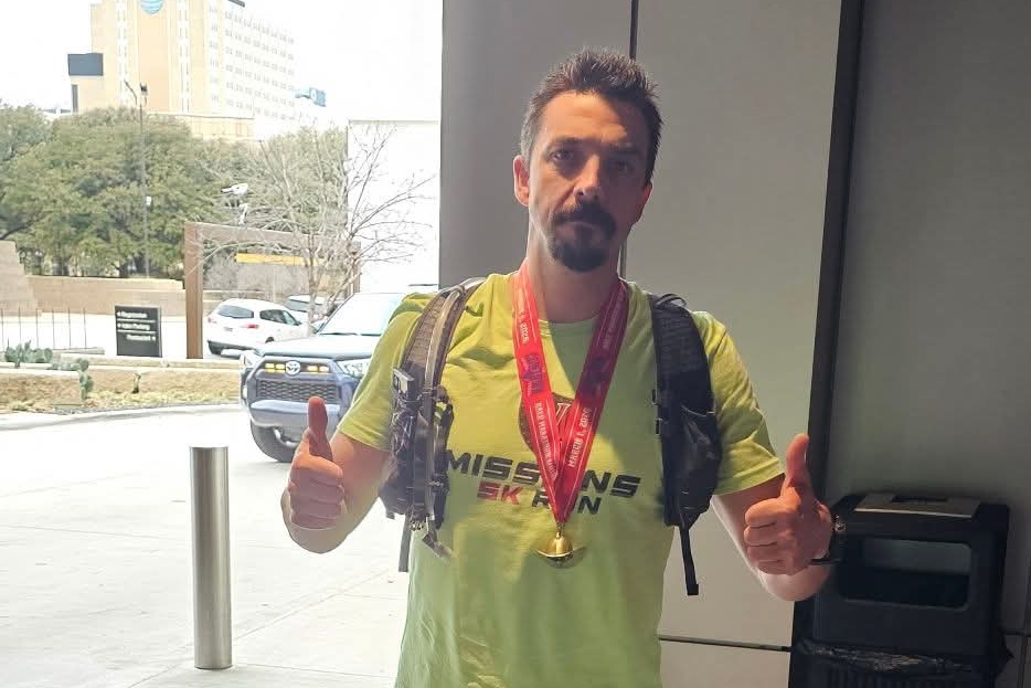 Man indoors wearing a neon yellow t‑shirt and backpack, with a red medal around his neck, giving thumbs up.