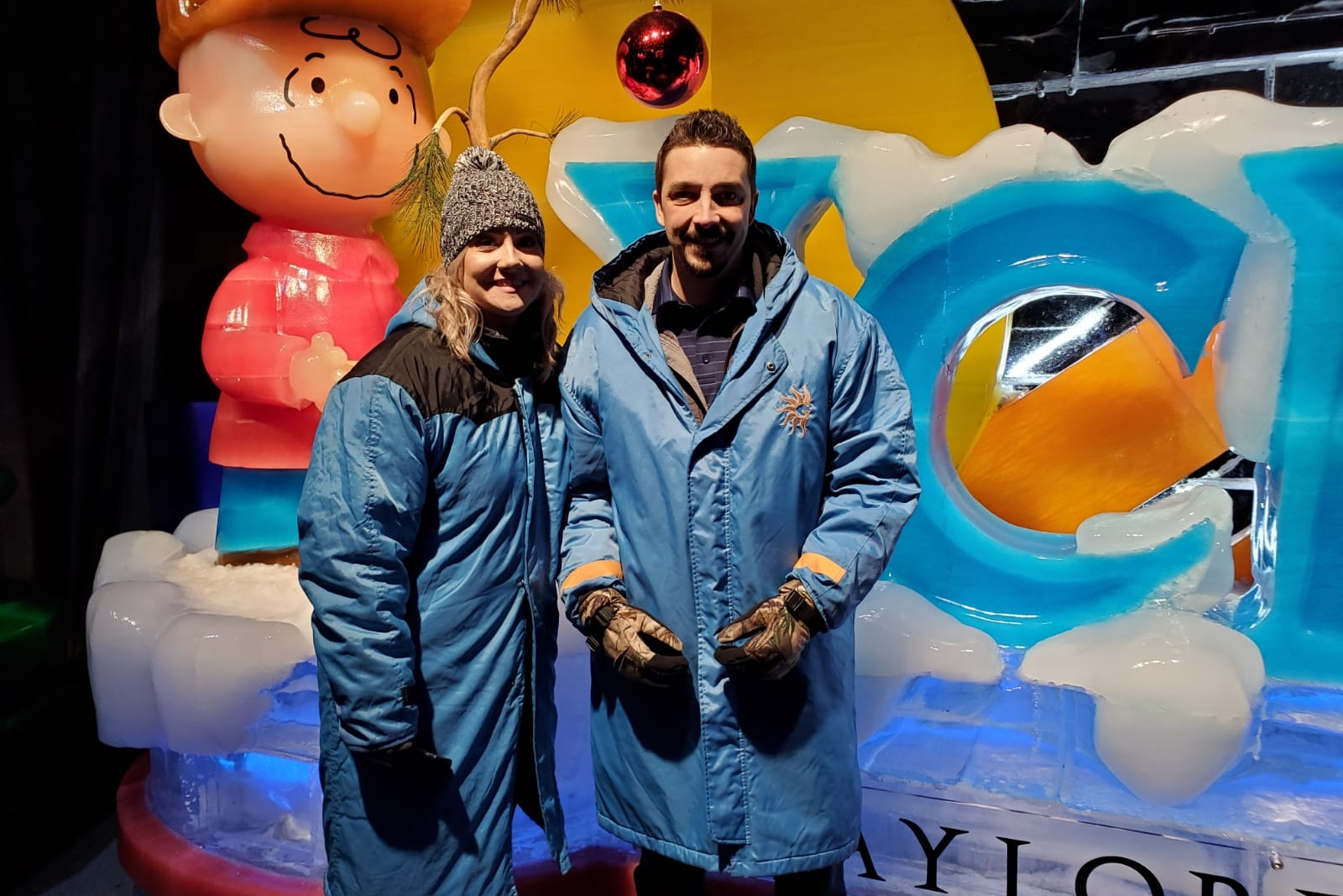 Two adults in blue winter jackets smile while posing in front of a colorful ice sculpture and a large cartoon statue behind them.