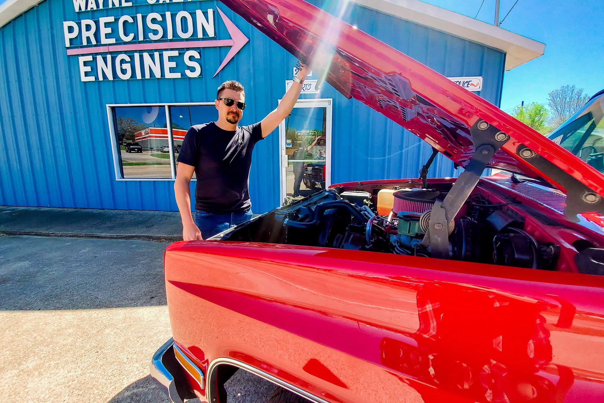 Man in a black shirt and sunglasses standing beside a red car with its hood open outside a blue shop labeled Precision Engines.