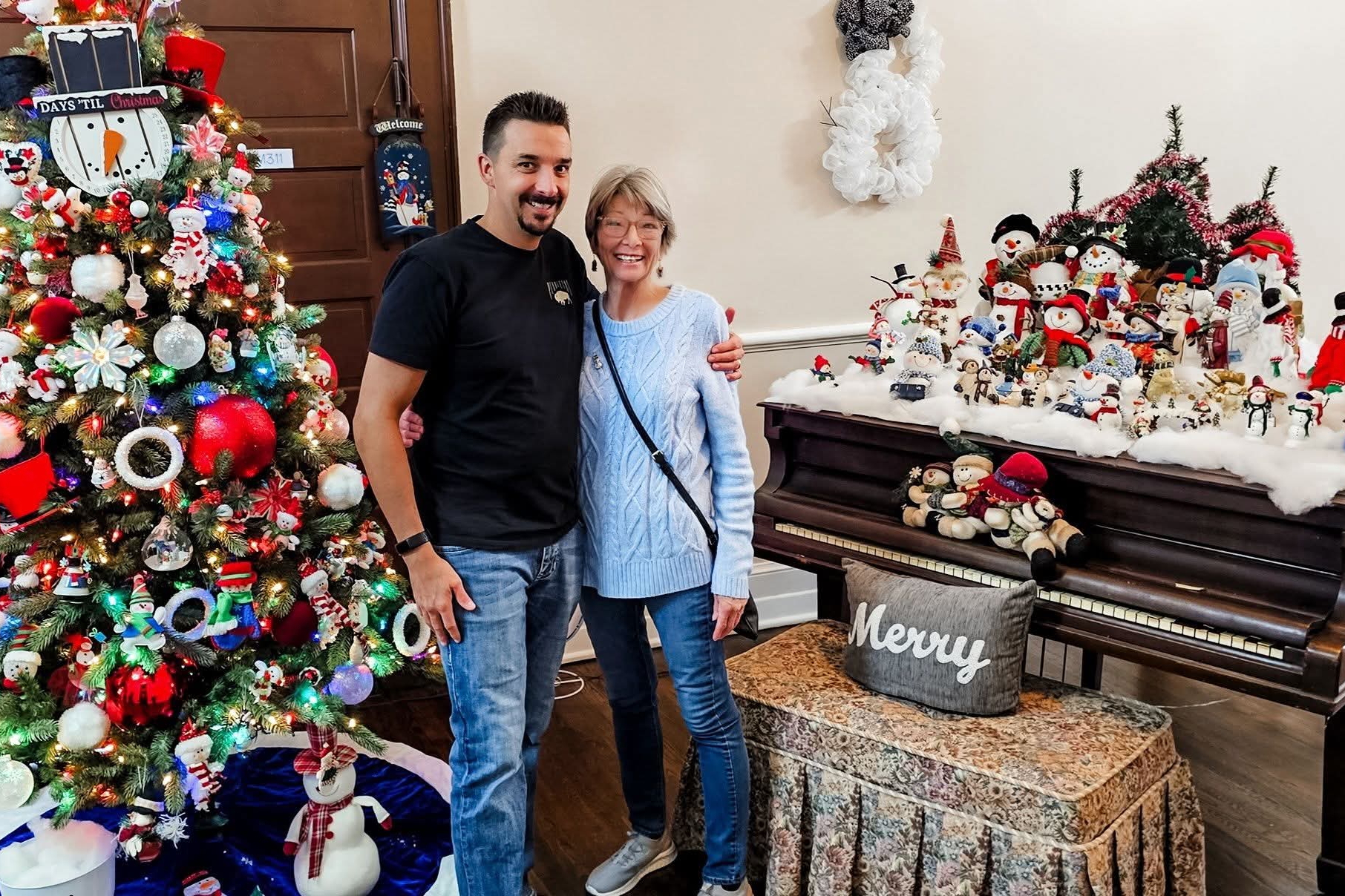 Smiling couple standing beside a decorated Christmas tree with colorful lights and ornaments, and a piano display of snowmen and holiday decor in the background.